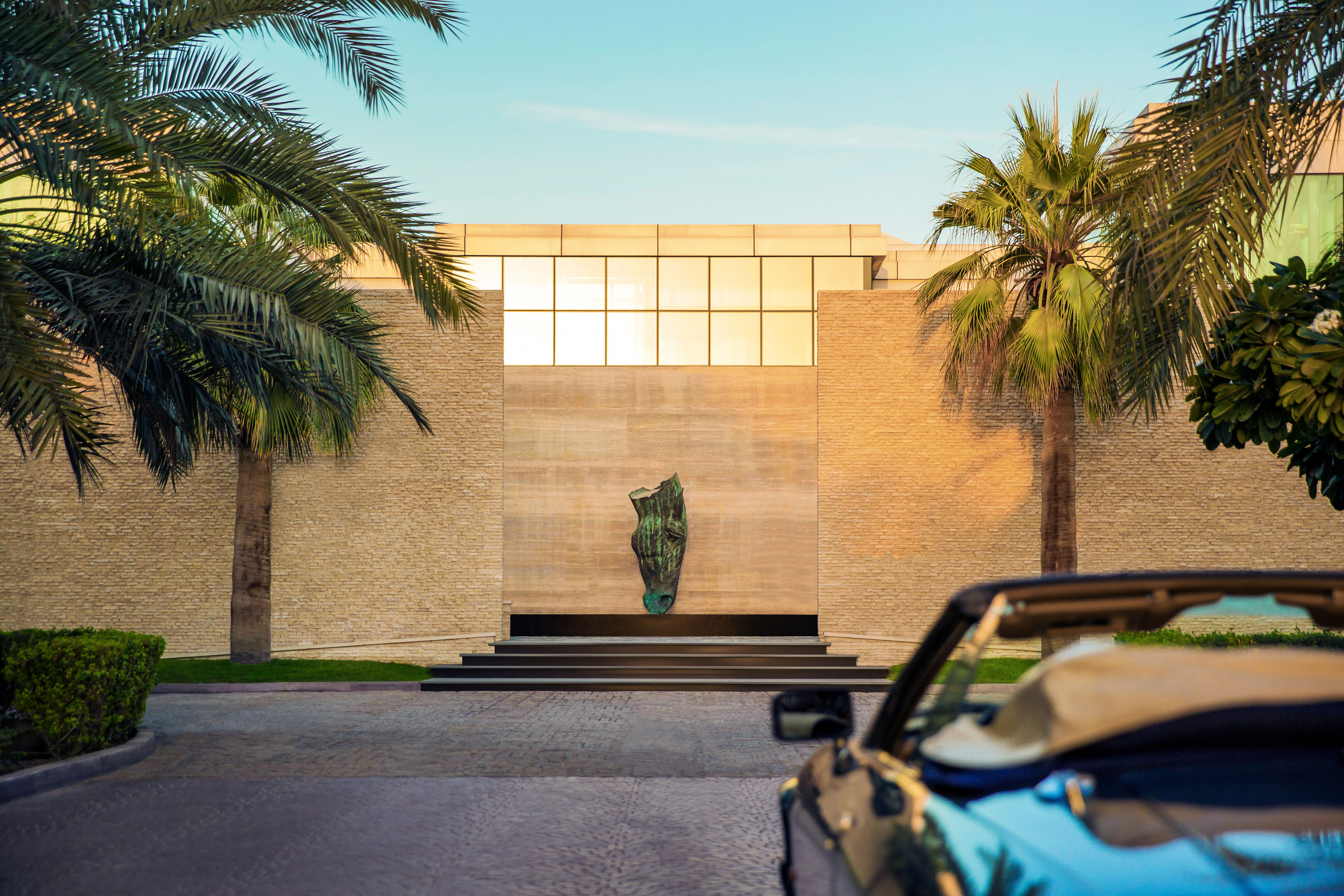Modern building entrance, green sculpture, palm trees, luxury convertible.