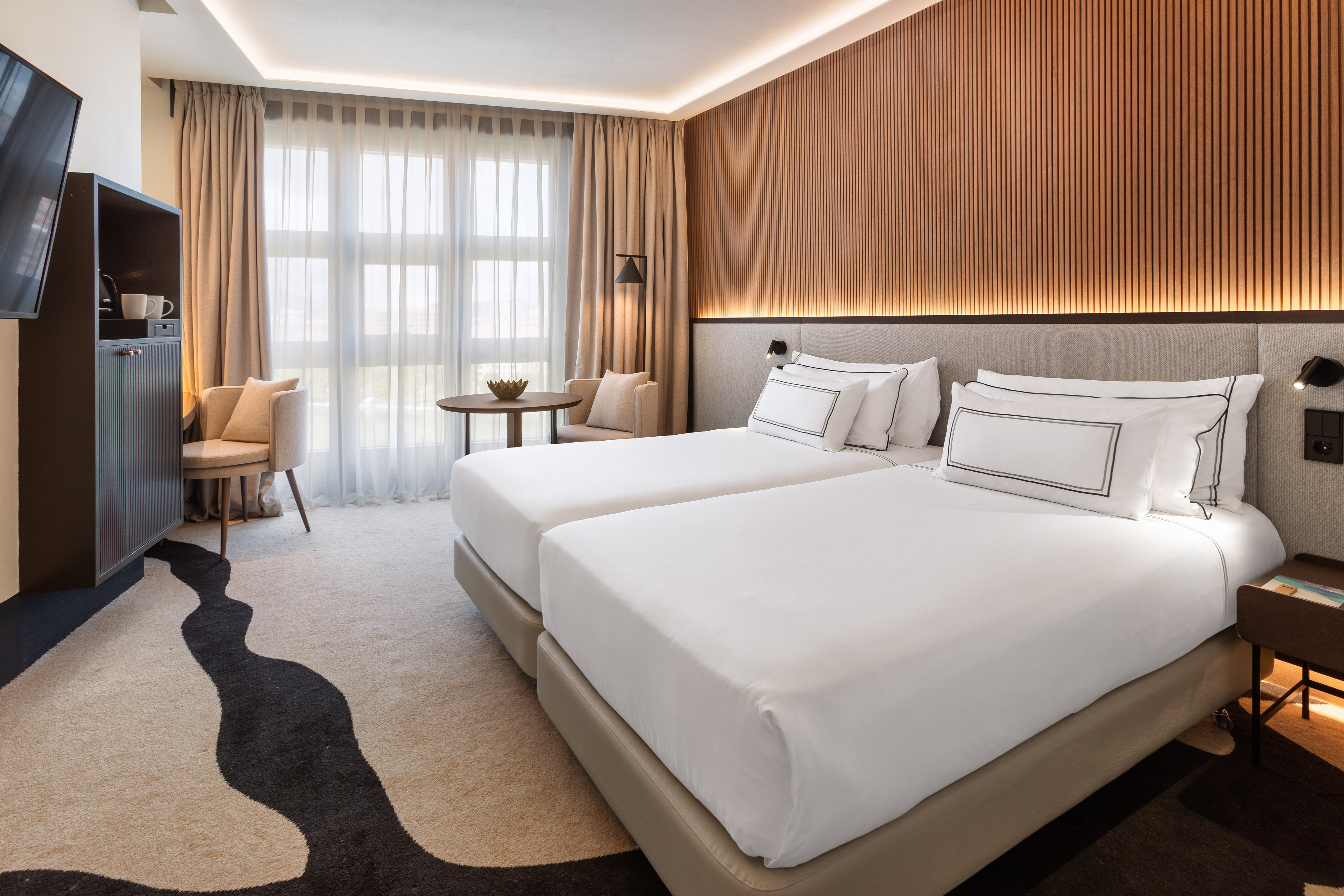 Modern hotel room with twin beds, wood wall decor, bright window, and patterned carpet.