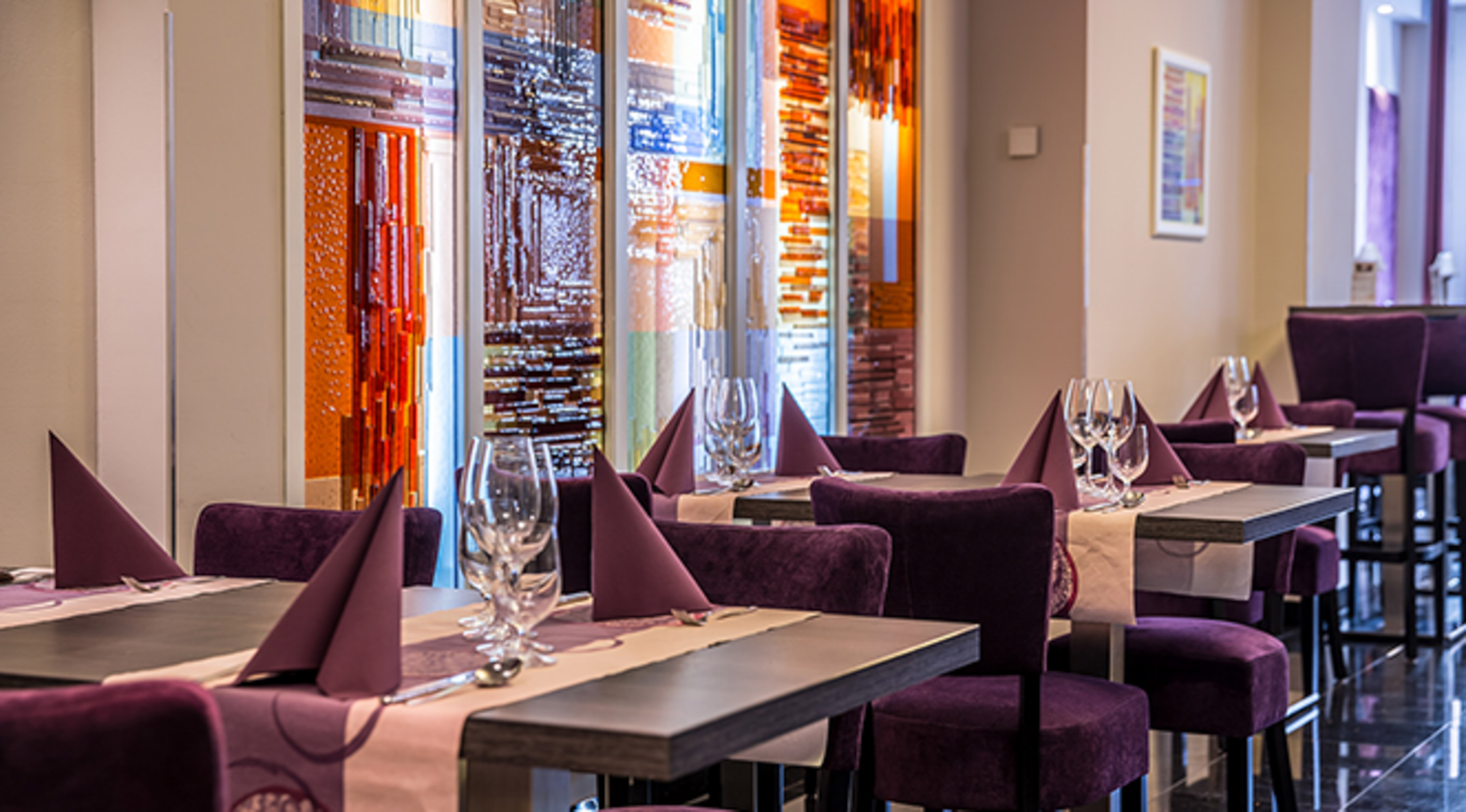 Modern restaurant interior with purple velvet chairs and colorful glass panels.