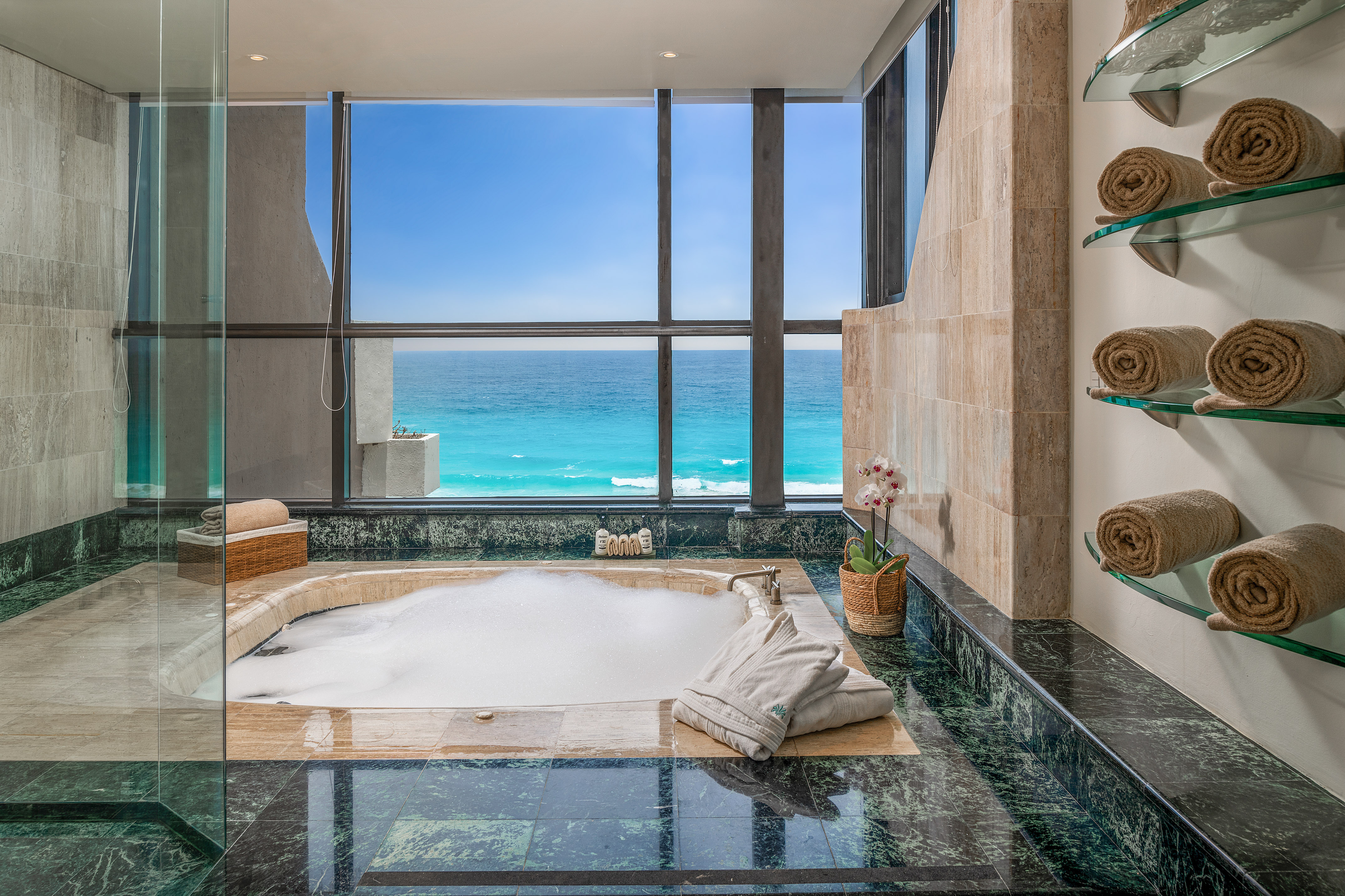Luxury ocean view bathroom with large windows, bubble bath, marble floor.