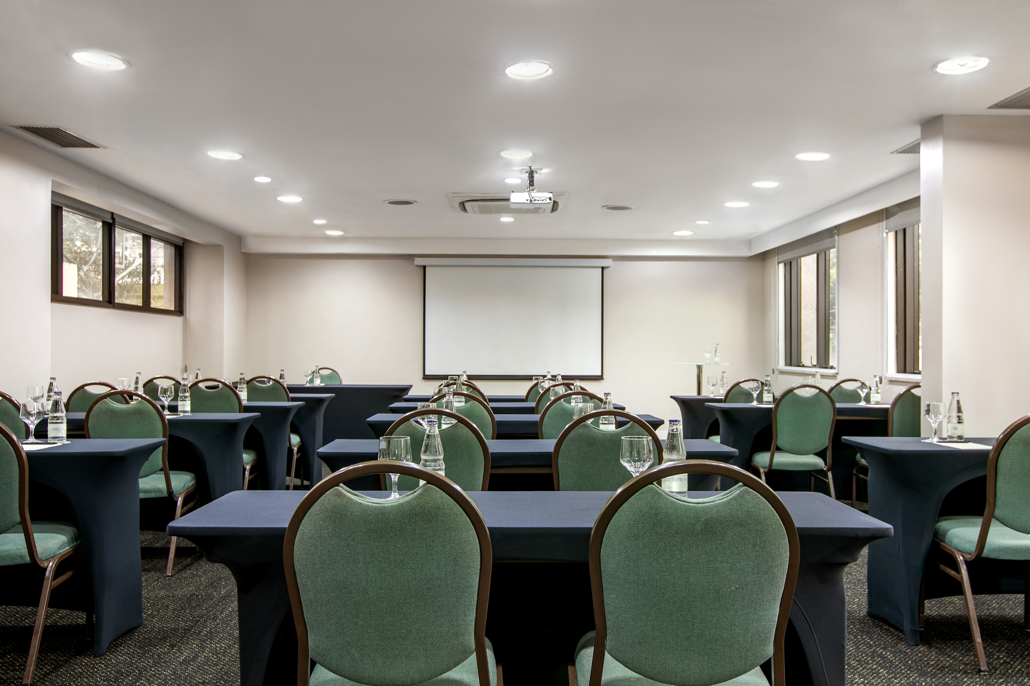 Empty conference room with green chairs, dark tables, projector, and screen.