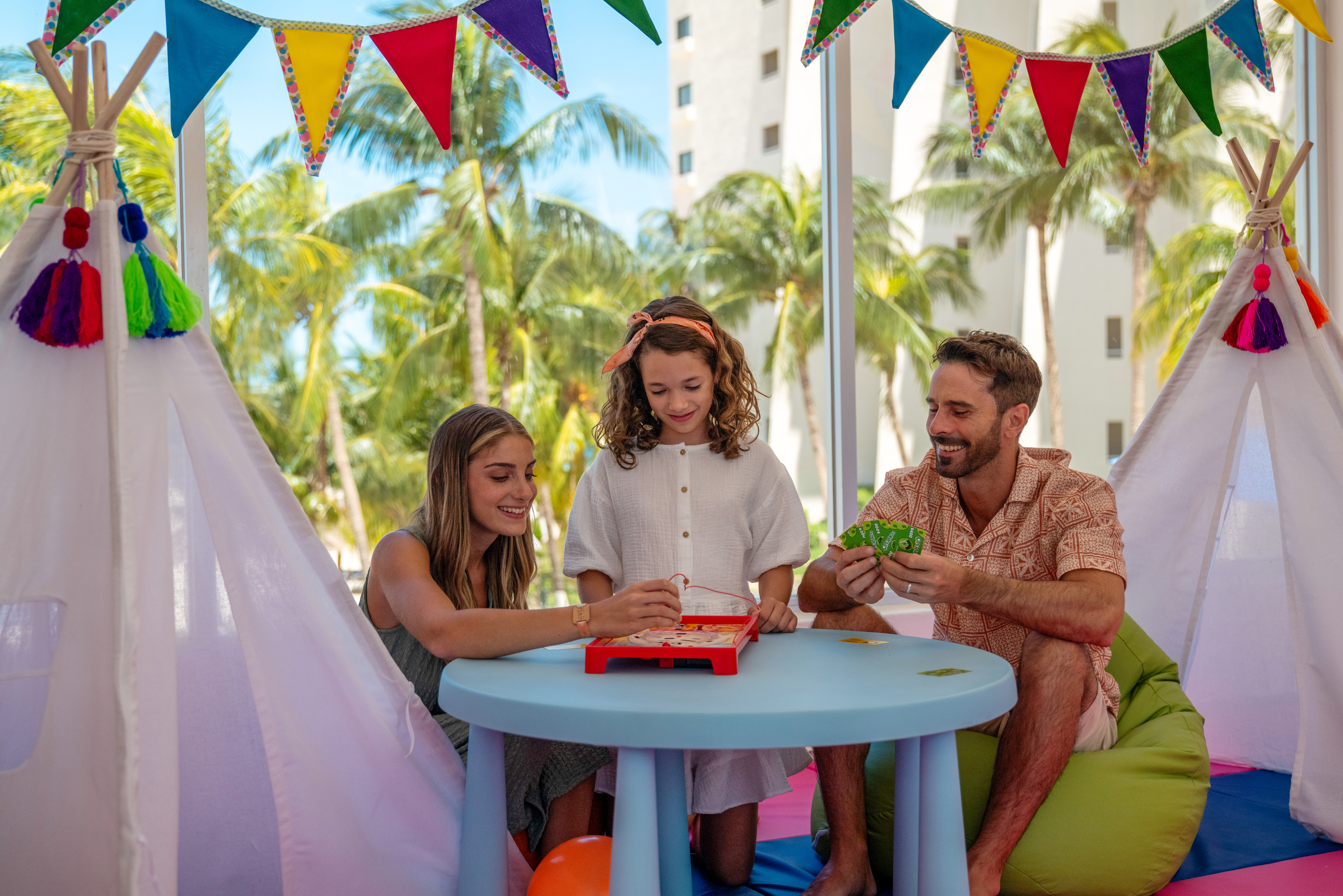 Family enjoying a board game in a colorful resort kids club with palm trees.