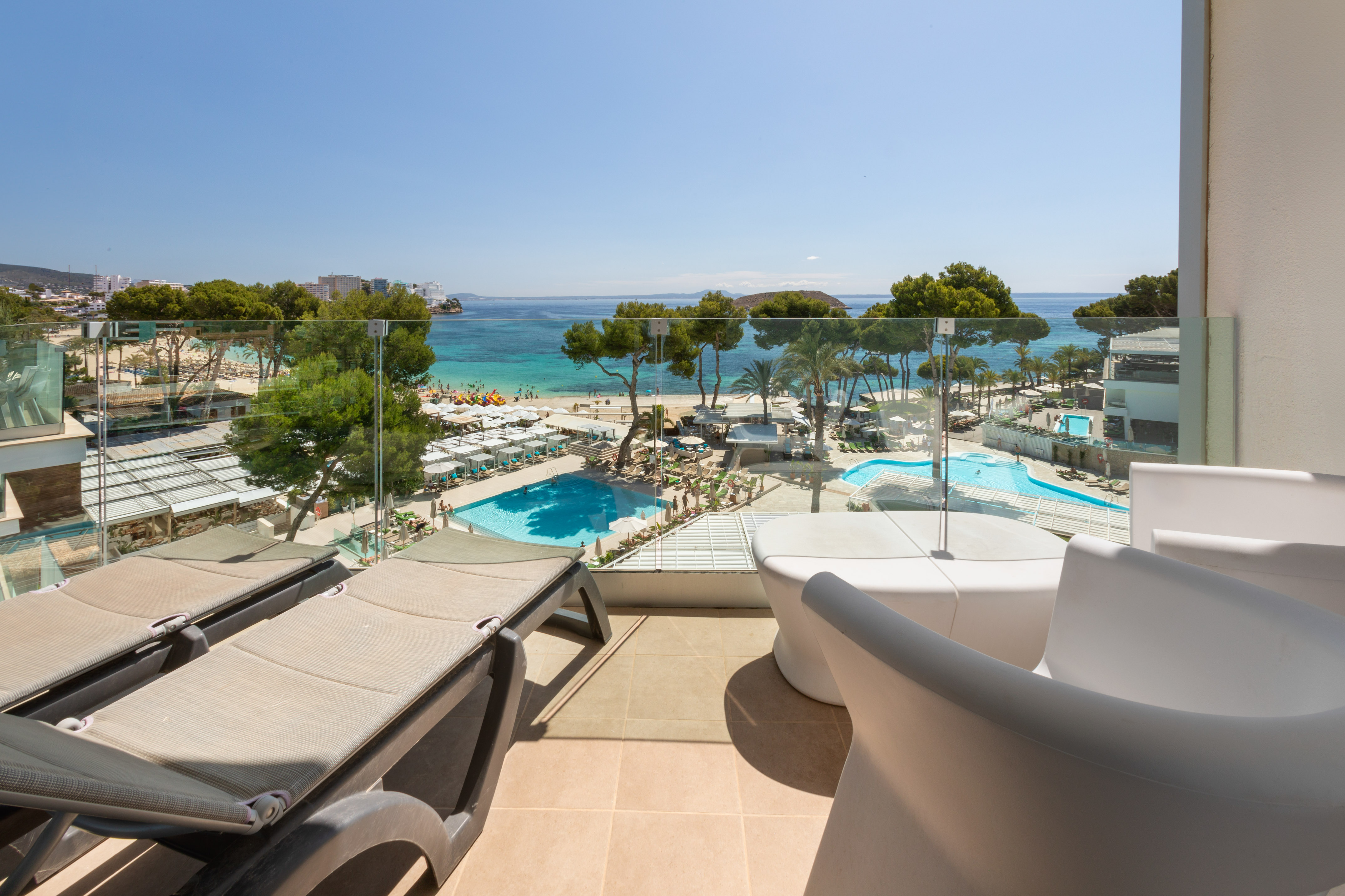 Sunny hotel balcony overlooking a sandy beach, blue ocean, and resort swimming pools.