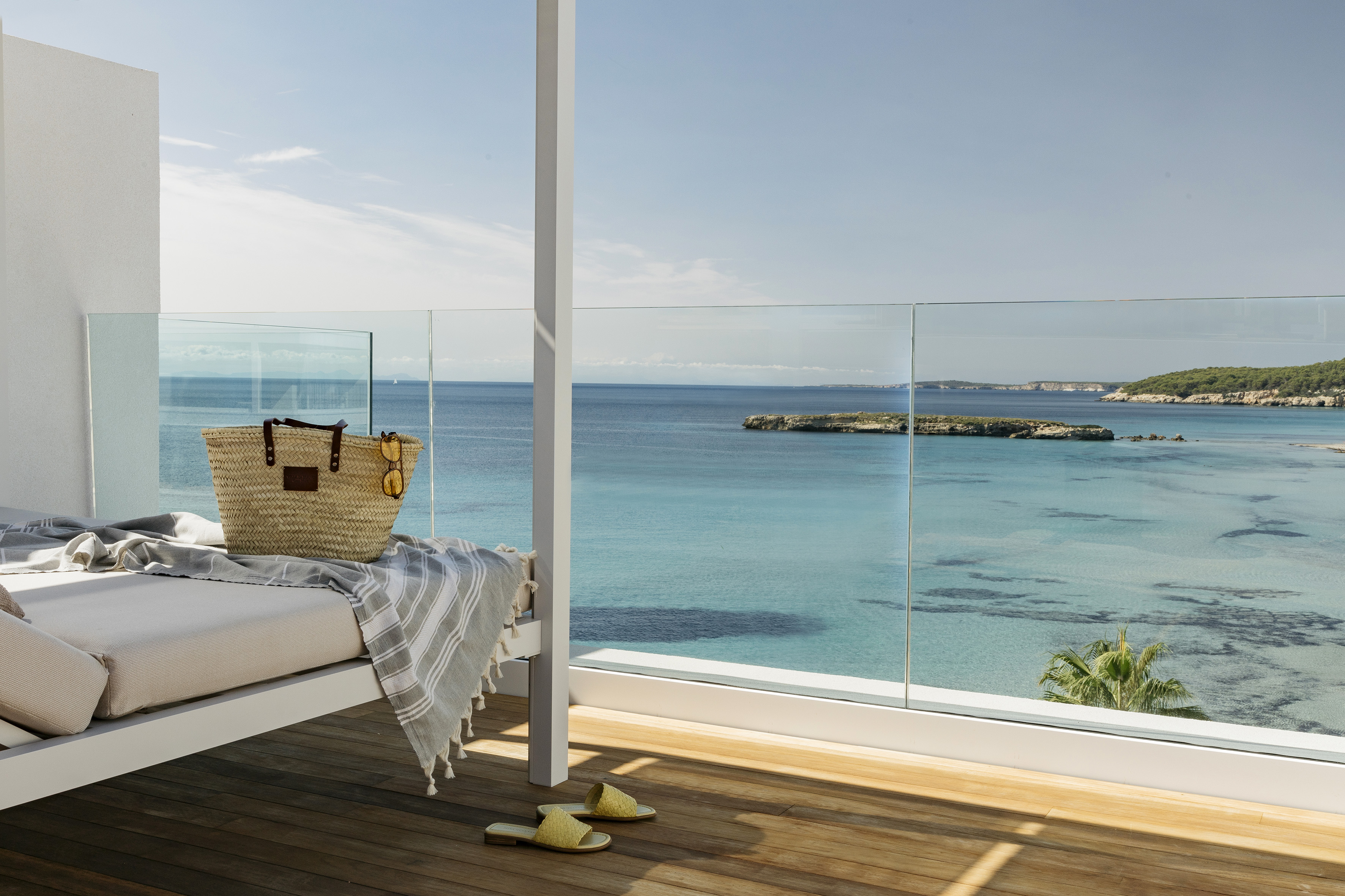 Beachfront balcony with daybed, straw bag, sandals, and turquoise sea.