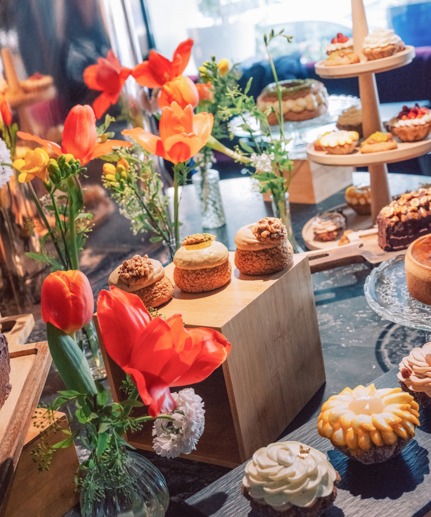 Gourmet dessert display with choux pastries, tarts, cakes, and red tulips.