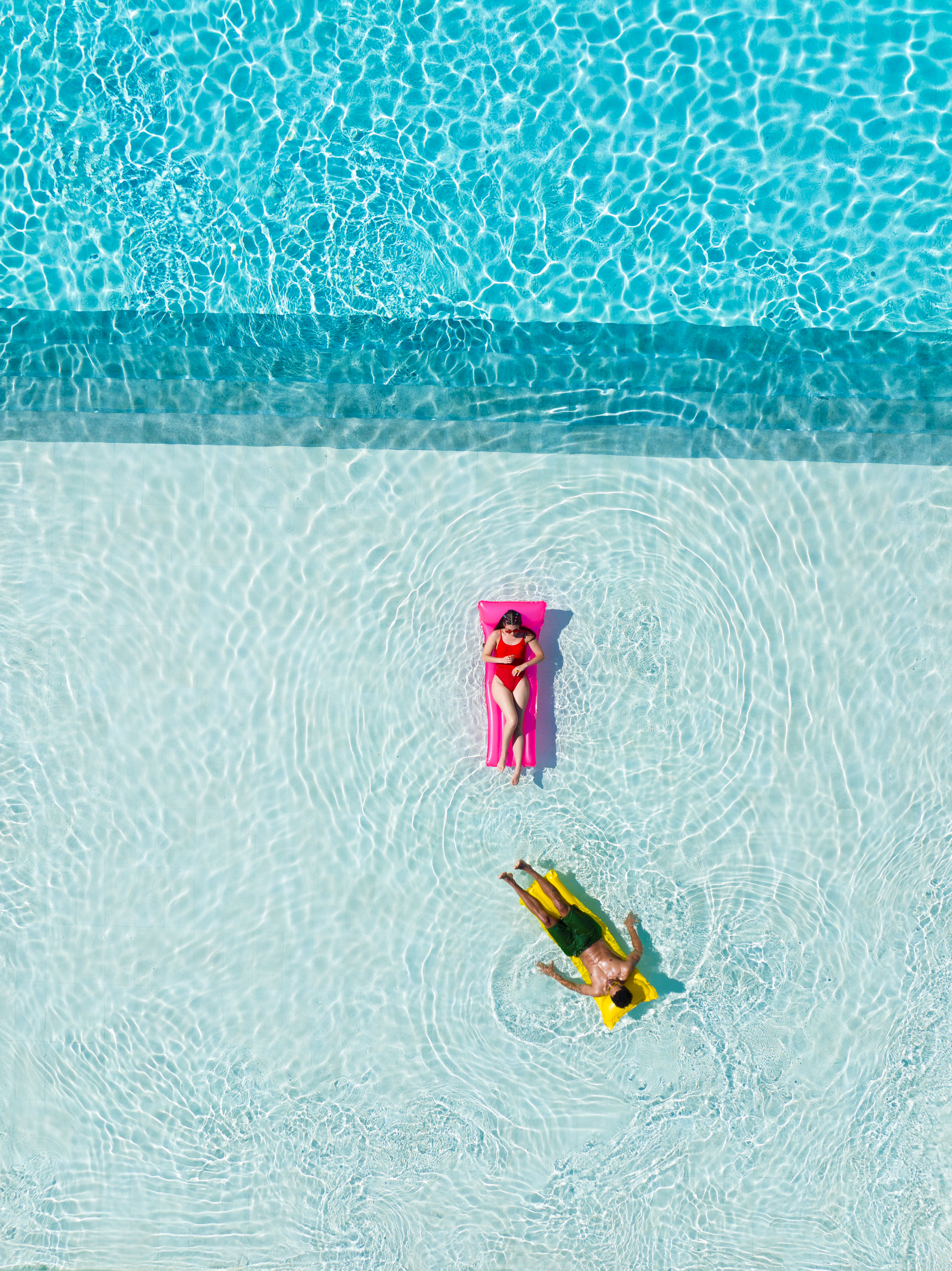 Couple floating on rafts in bright blue swimming pool, summer relaxation.