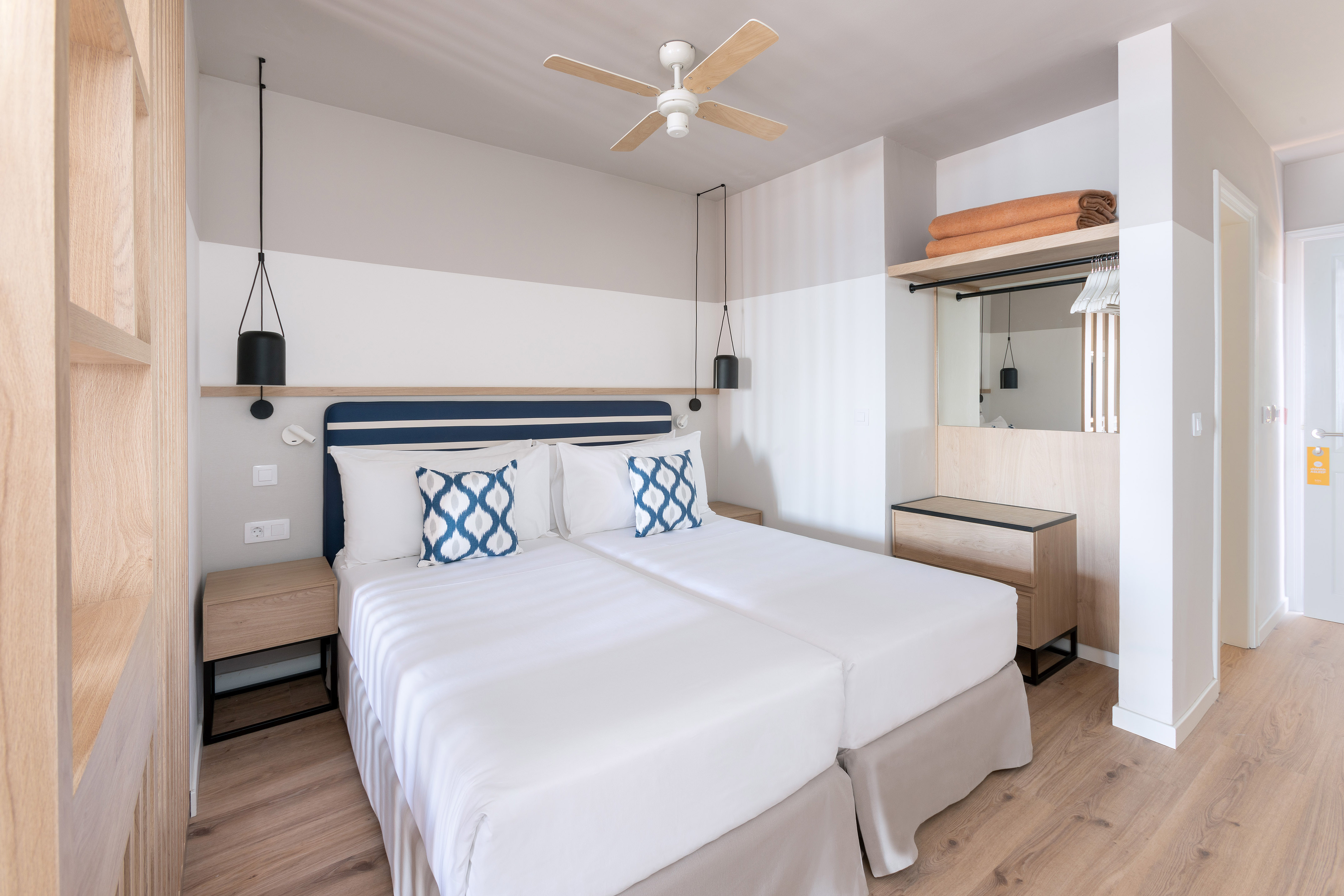Bright hotel room with twin beds, blue pillows, wooden floor, and ceiling fan.