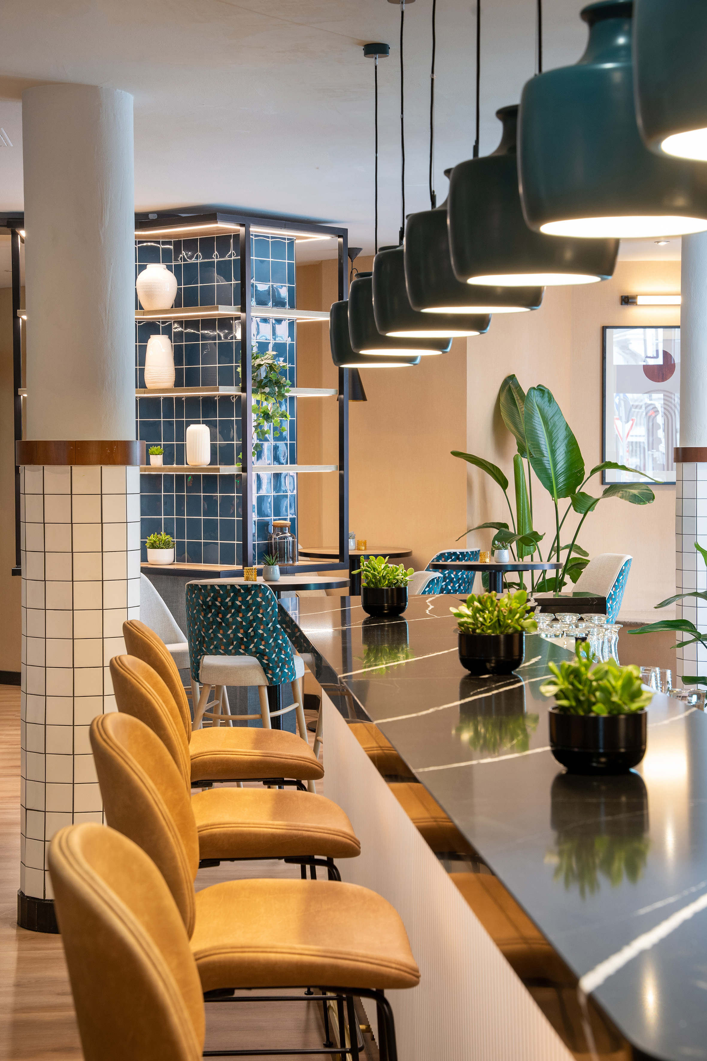 Modern bar with black counter, camel stools, teal pendant lights, and plants.