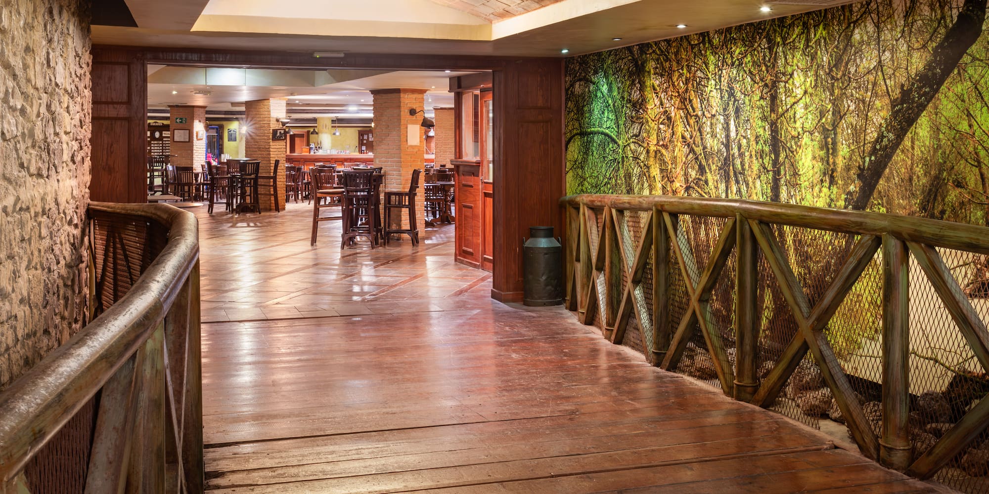 Rustic restaurant with wooden bridge, forest mural, and dining tables.