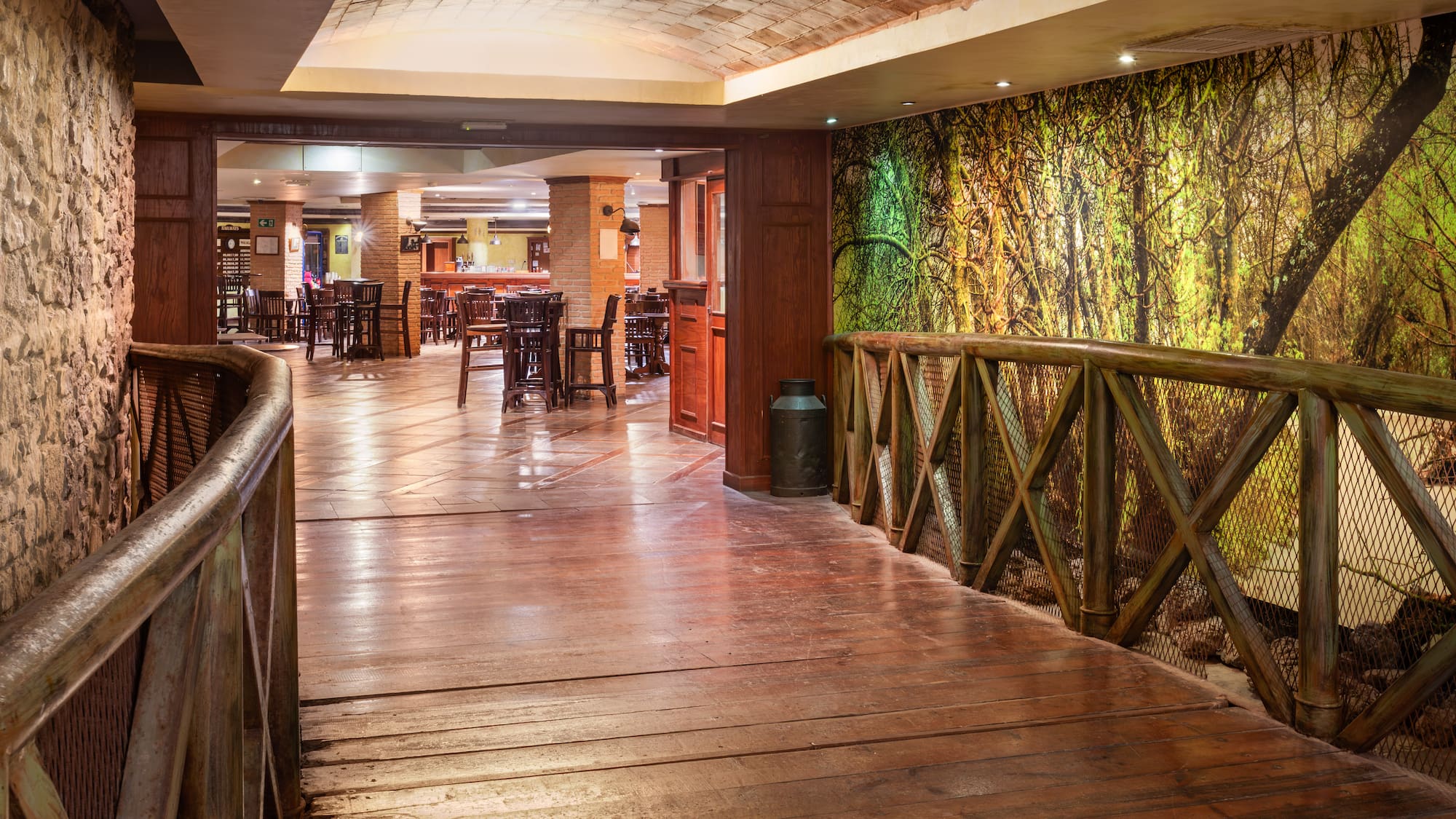 Rustic restaurant with wooden bridge, forest mural, and dining tables.