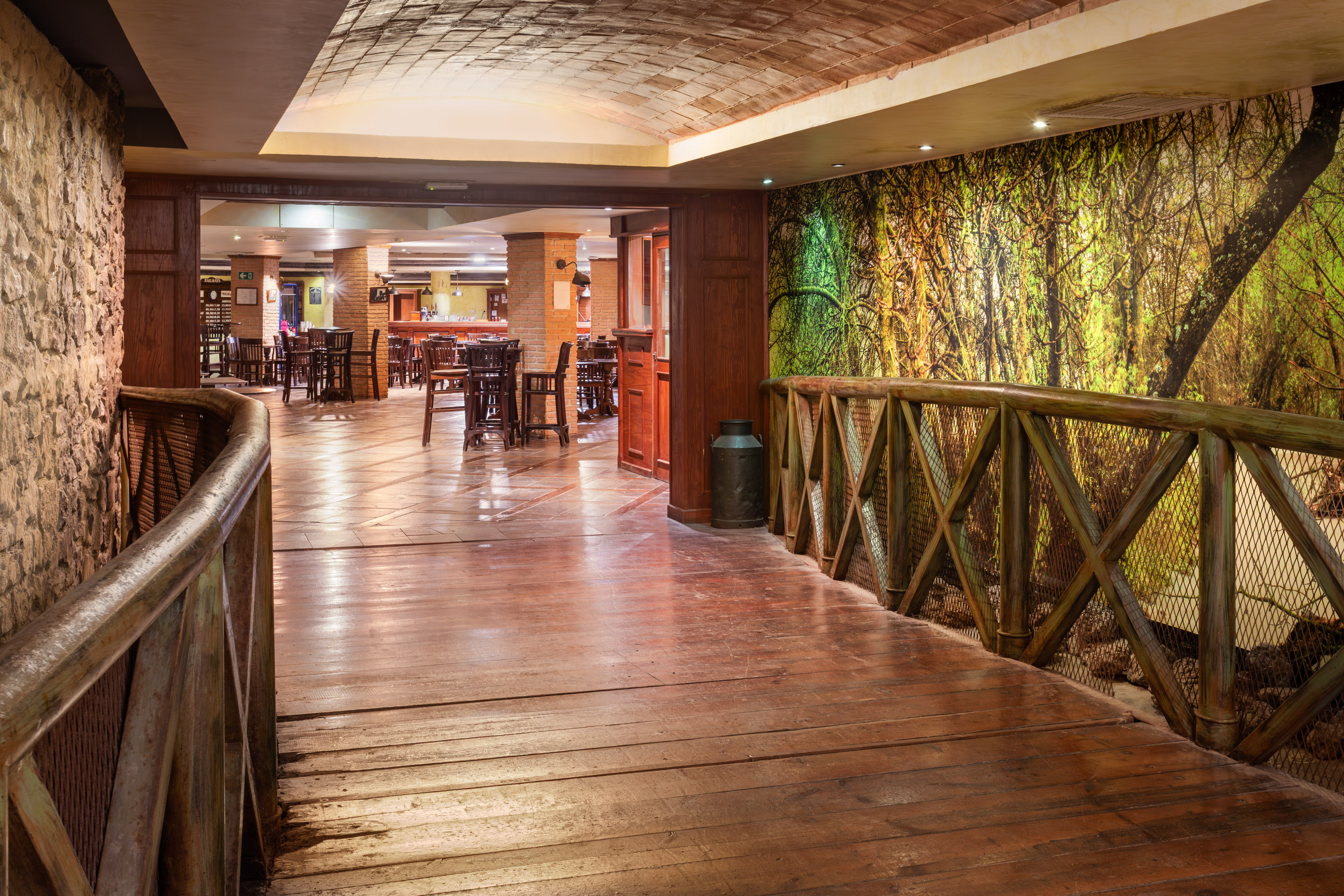 Rustic restaurant with wooden bridge, forest mural, and dining tables.