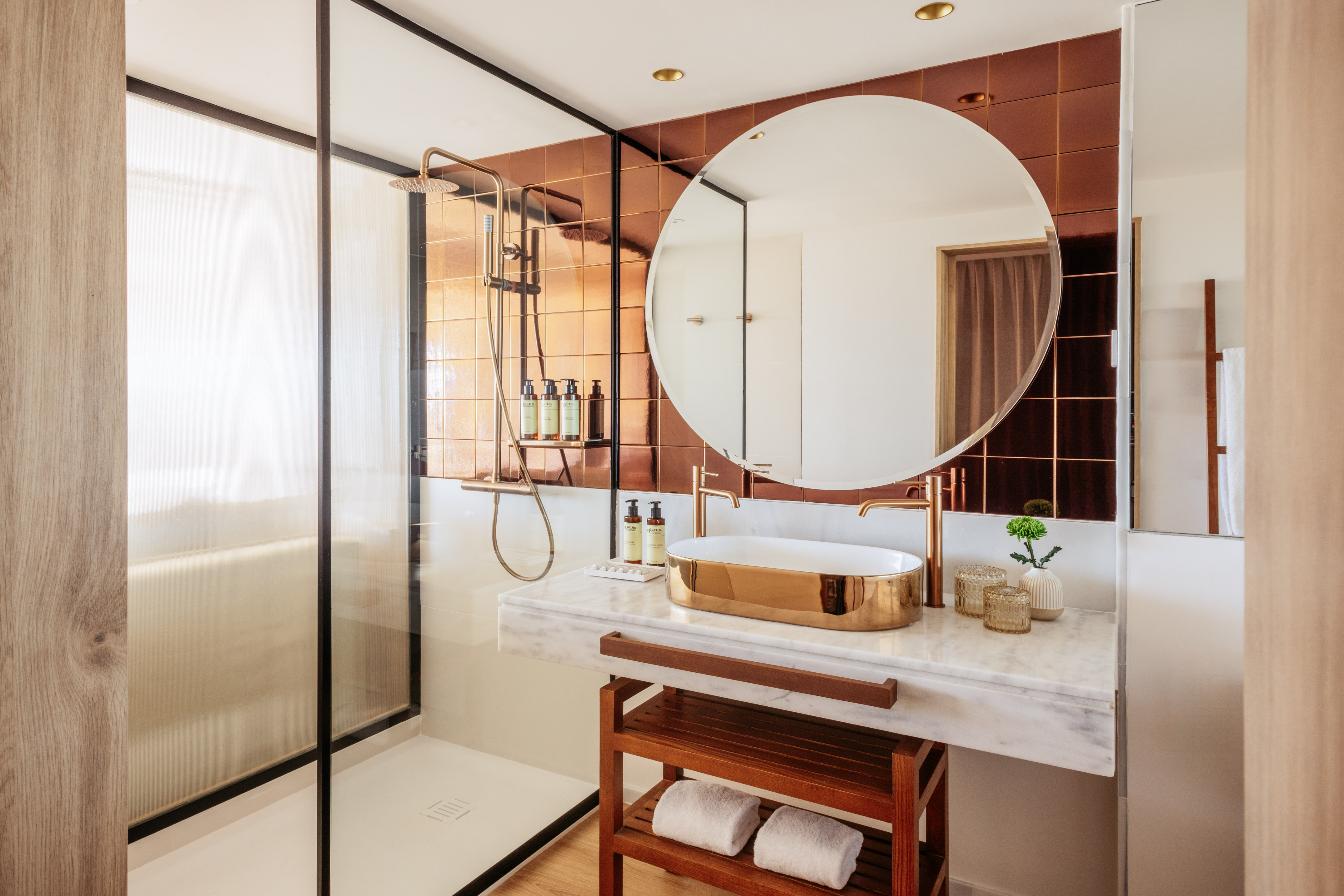 Modern bathroom with gold vessel sink, marble vanity, copper wall tiles, glass shower.