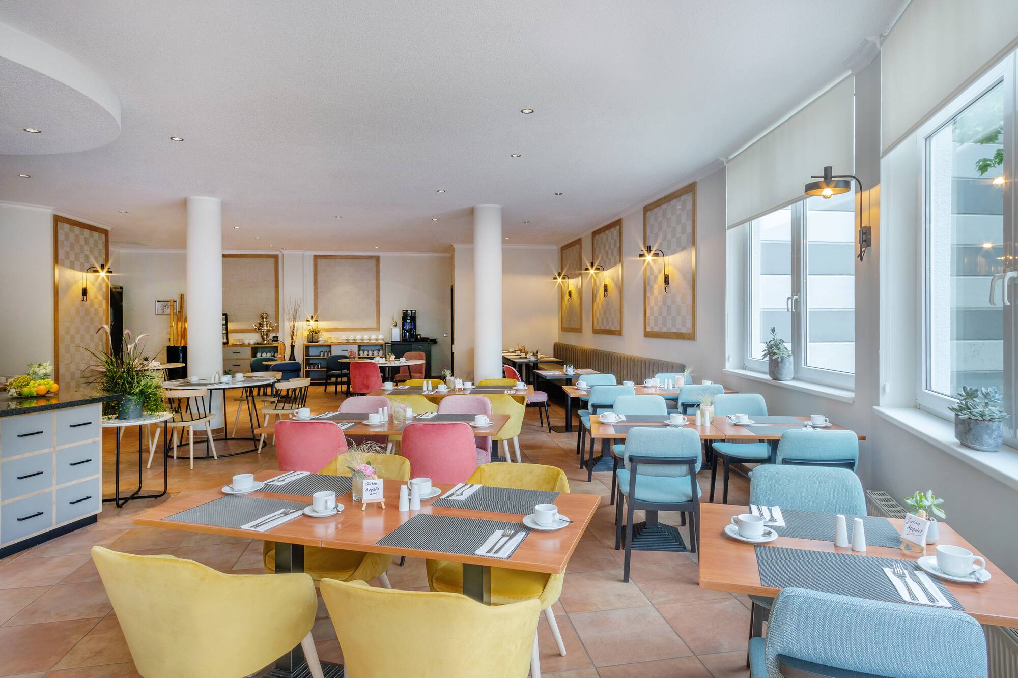 Bright hotel breakfast room with colorful chairs, set tables, and buffet area.