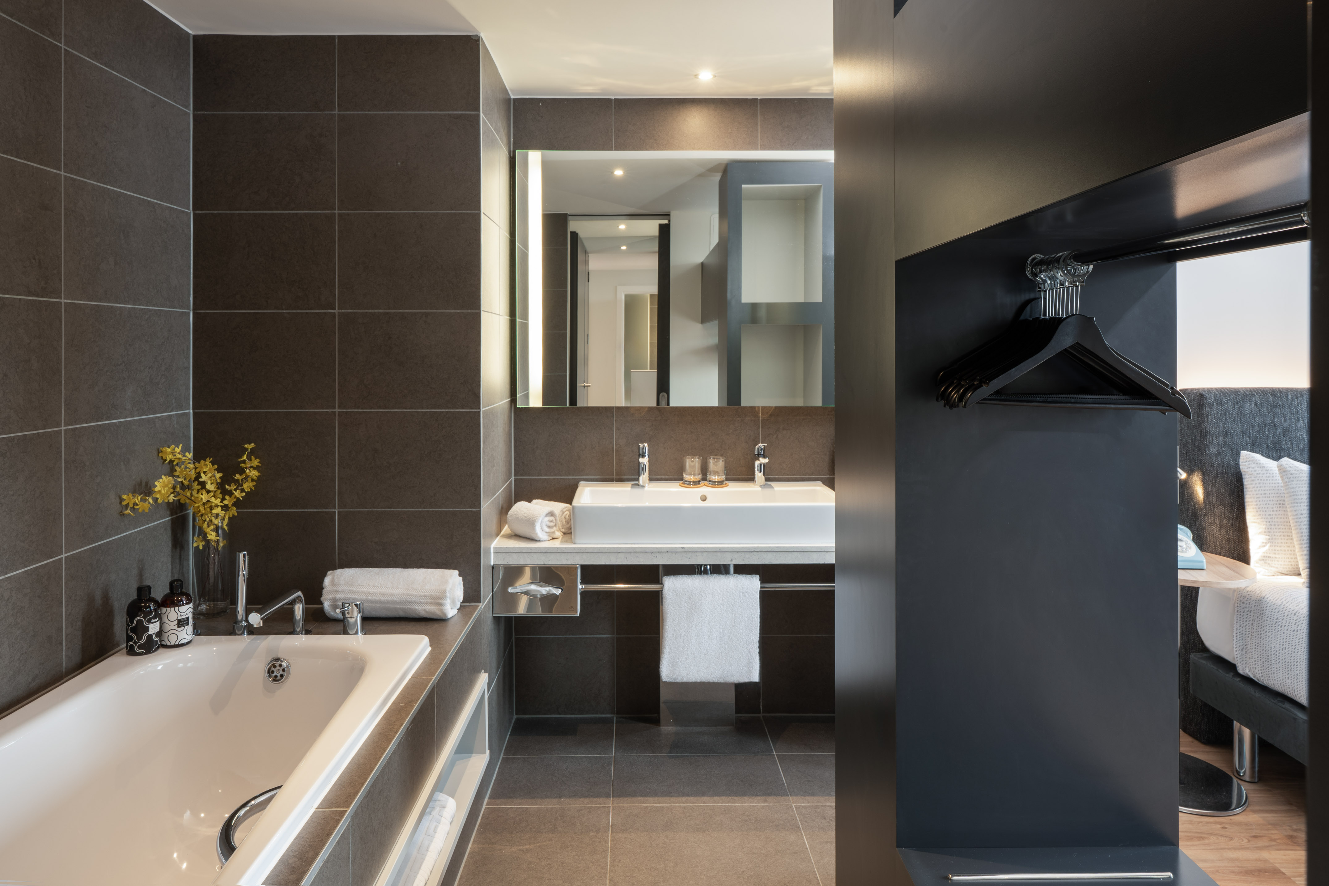 Modern hotel bathroom with dark tiles, bathtub, double sink, and bedroom.