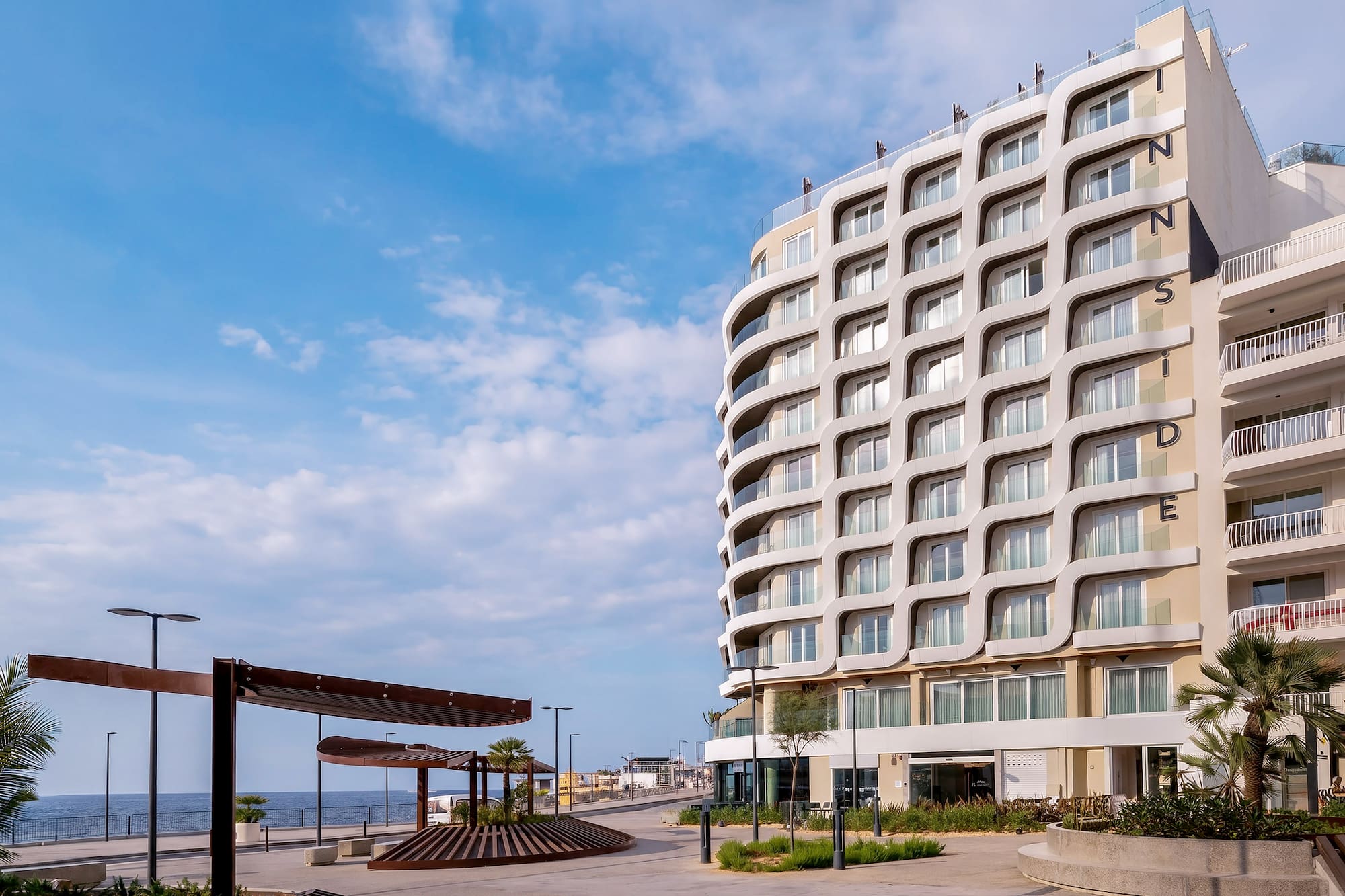 INNSiDE hotel with wavy facade overlooking a sunny seaside promenade.