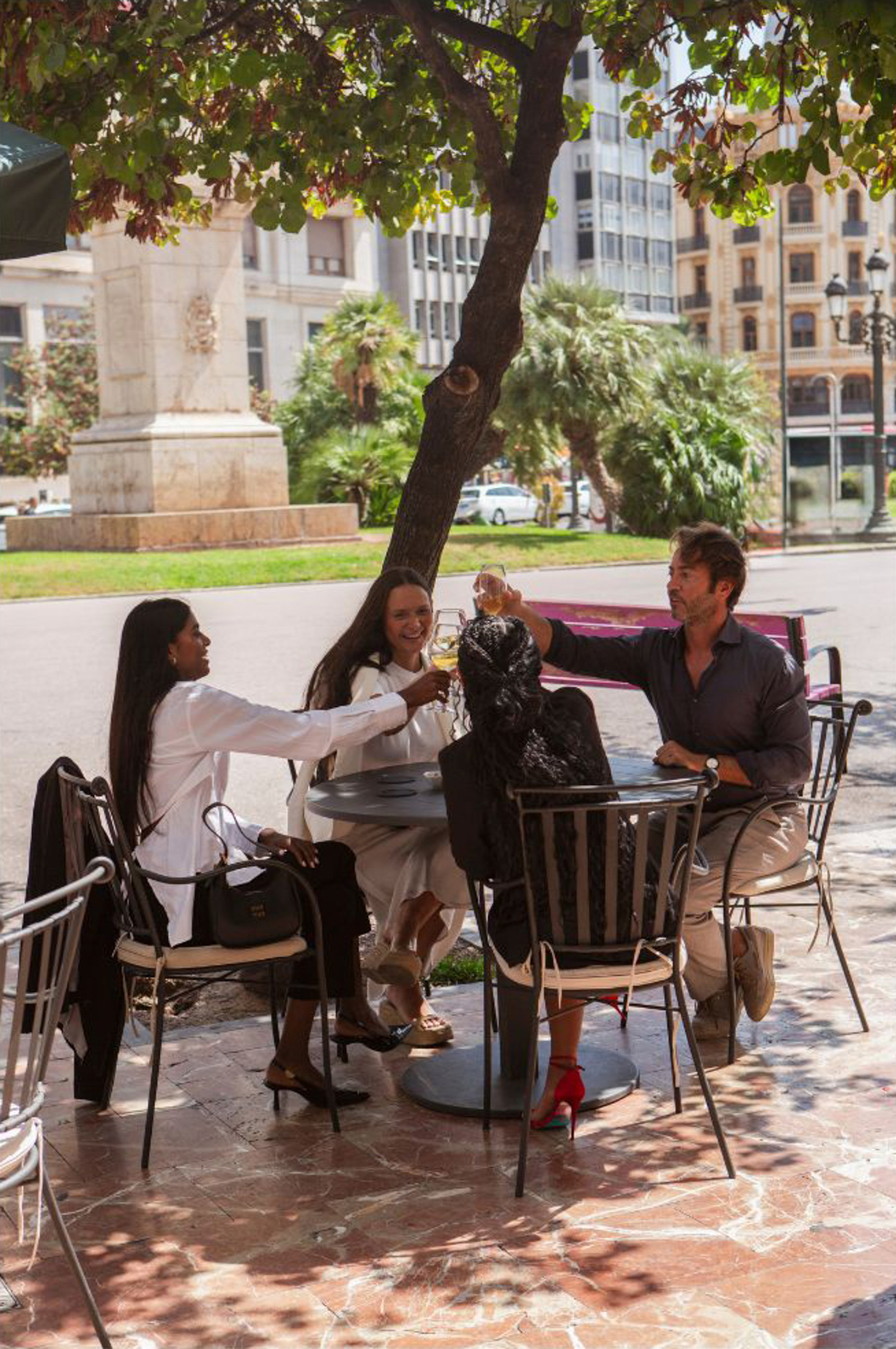 People toasting drinks at outdoor cafe table, city background, under tree shade.