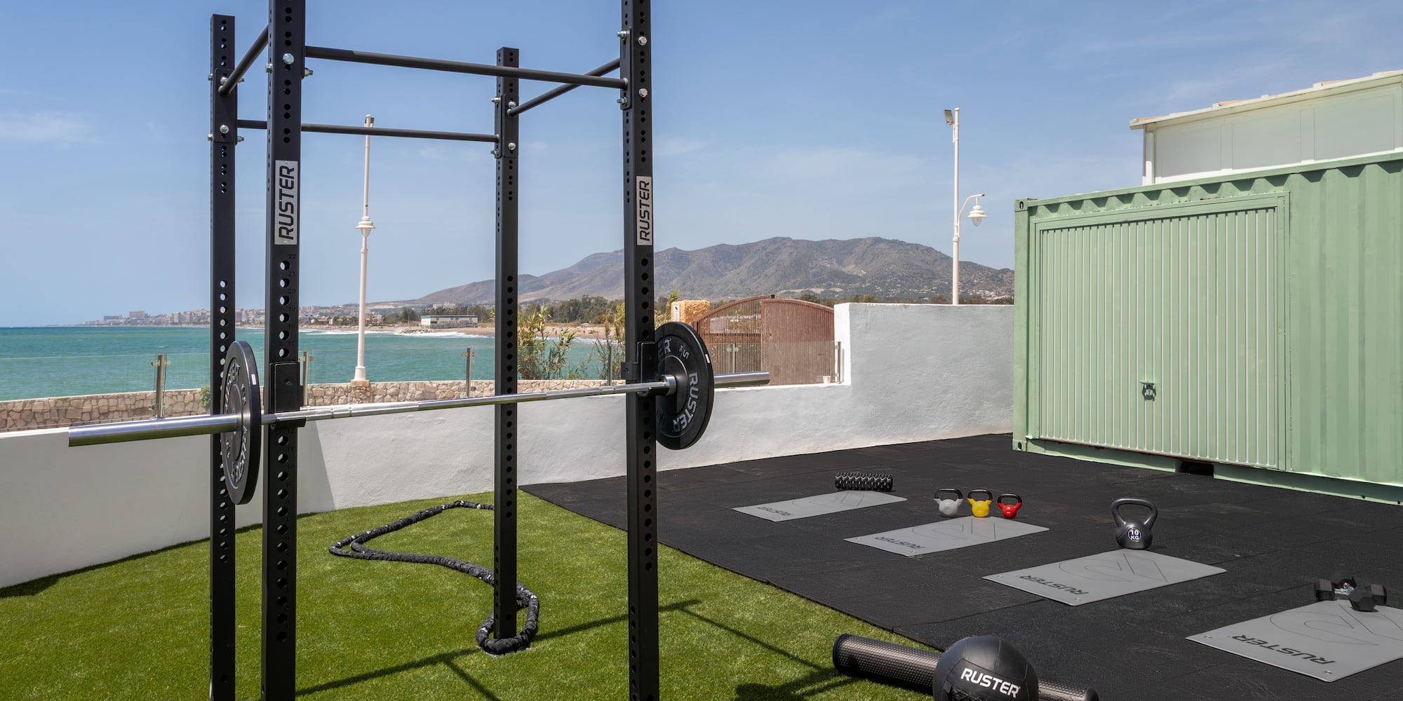 Ruster outdoor gym by sea: power rack, barbell, kettlebells, and exercise mats.
