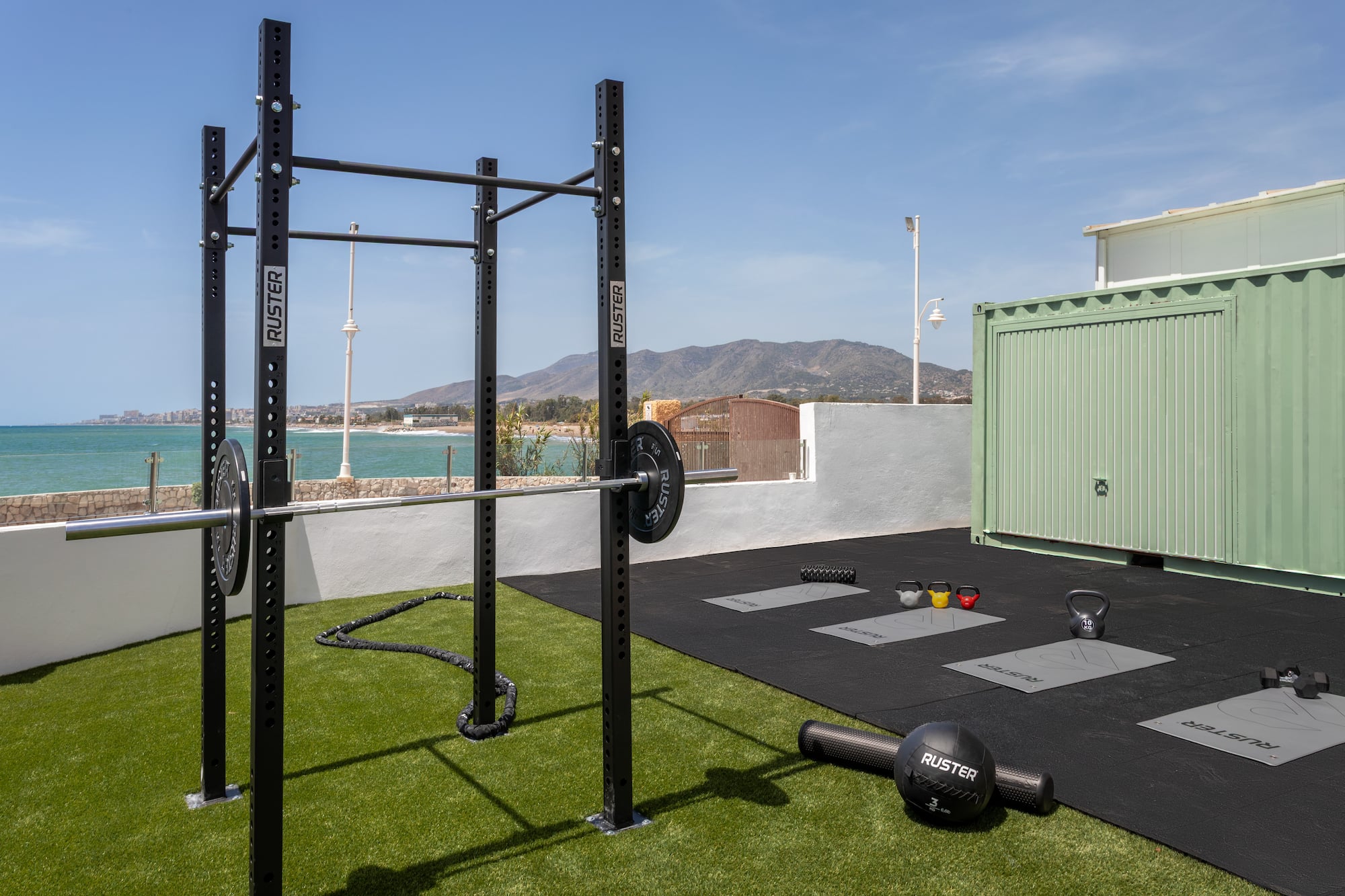 Gimnasio exterior Ruster con rack, pesas y kettlebells frente al mar.