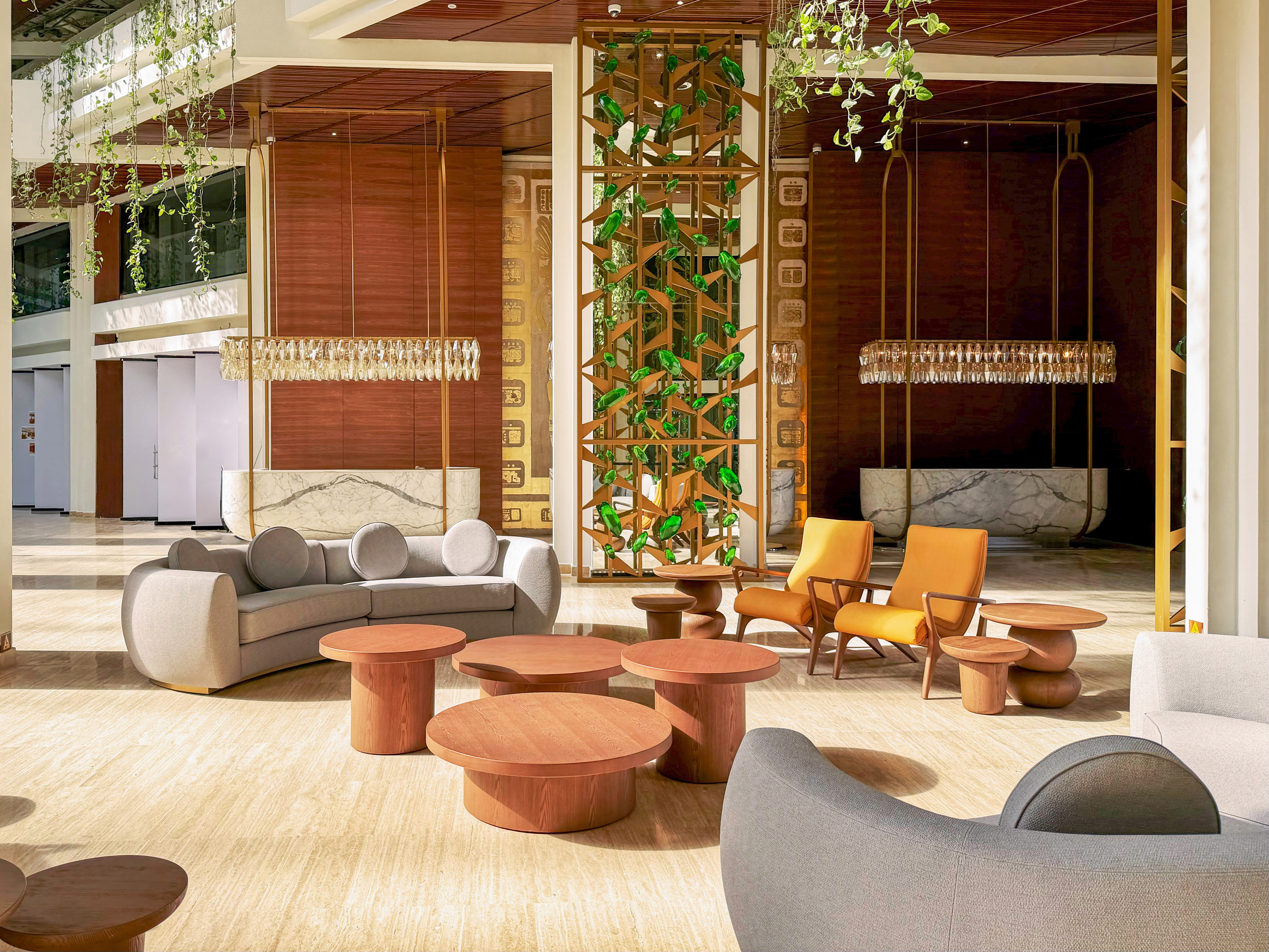 Modern hotel lobby lounge with sofa, orange chairs, wood tables, and green plants.