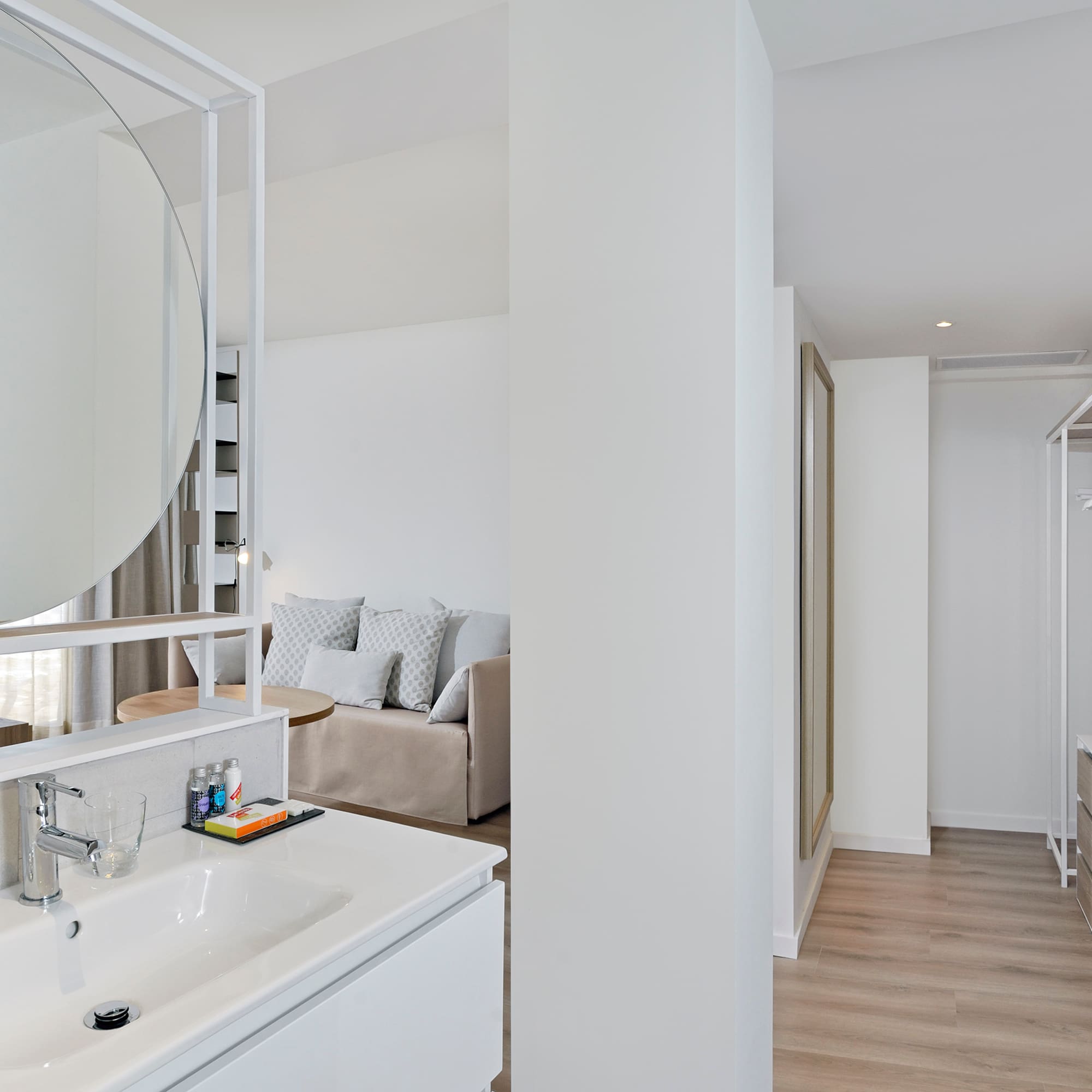 Modern hotel room: bathroom vanity, sofa area, open wardrobe.