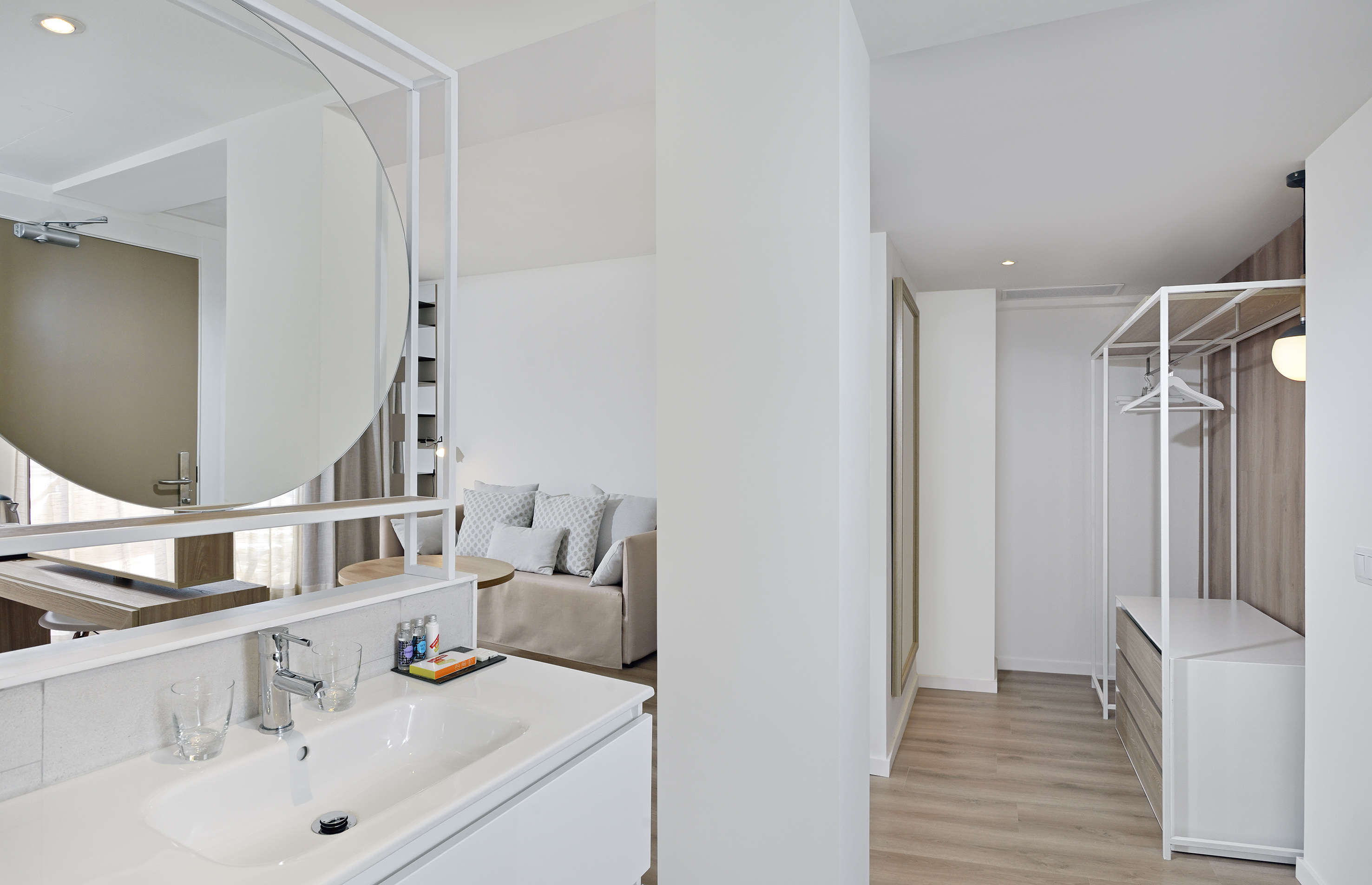 Modern hotel room: bathroom vanity, sofa area, open wardrobe.