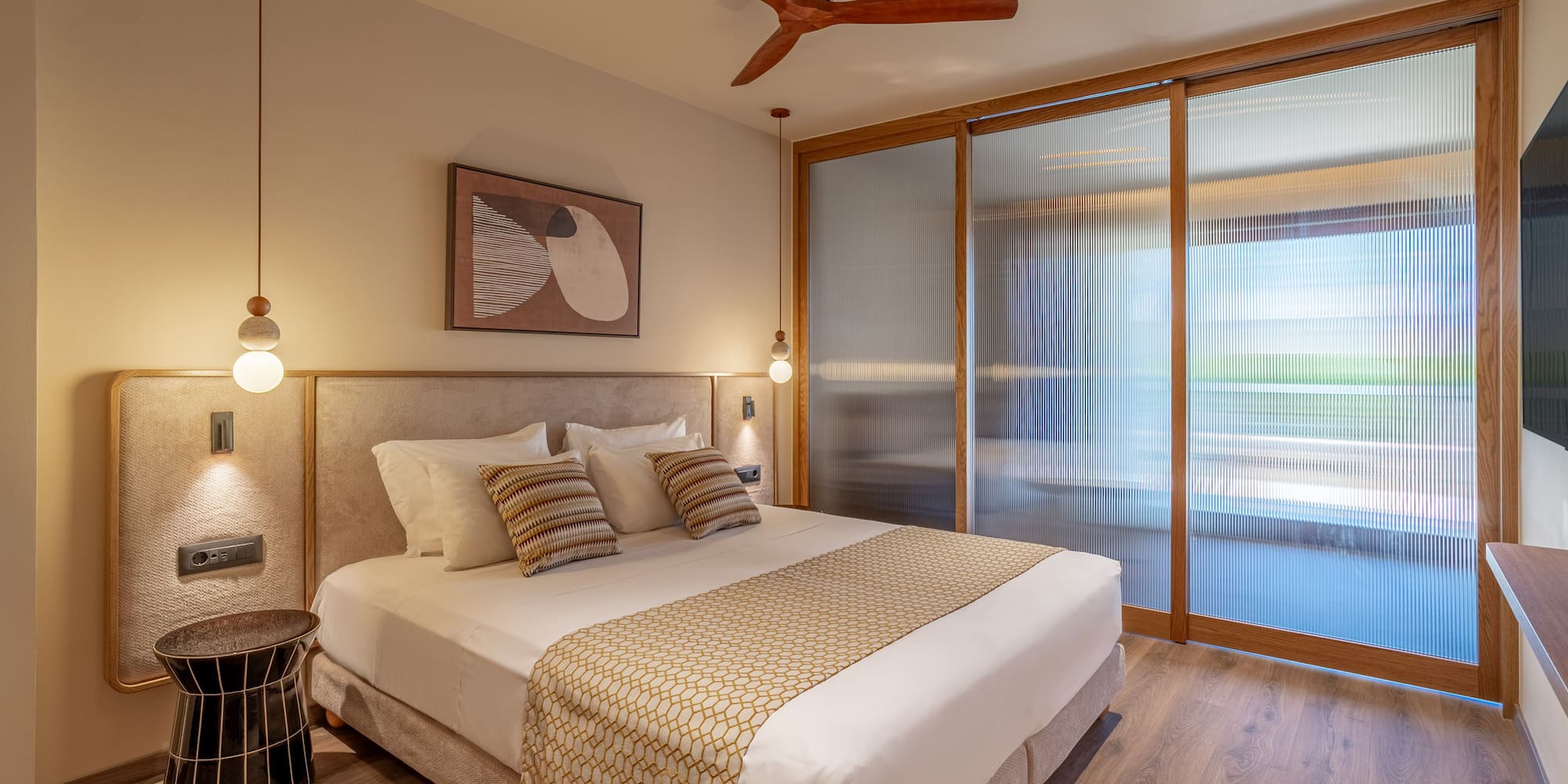 Modern hotel room with king bed, wooden ceiling fan, and fluted glass doors.