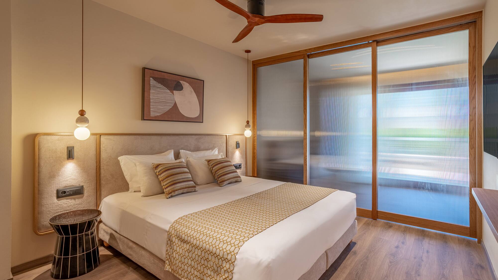 Modern hotel room with king bed, wooden ceiling fan, and fluted glass doors.