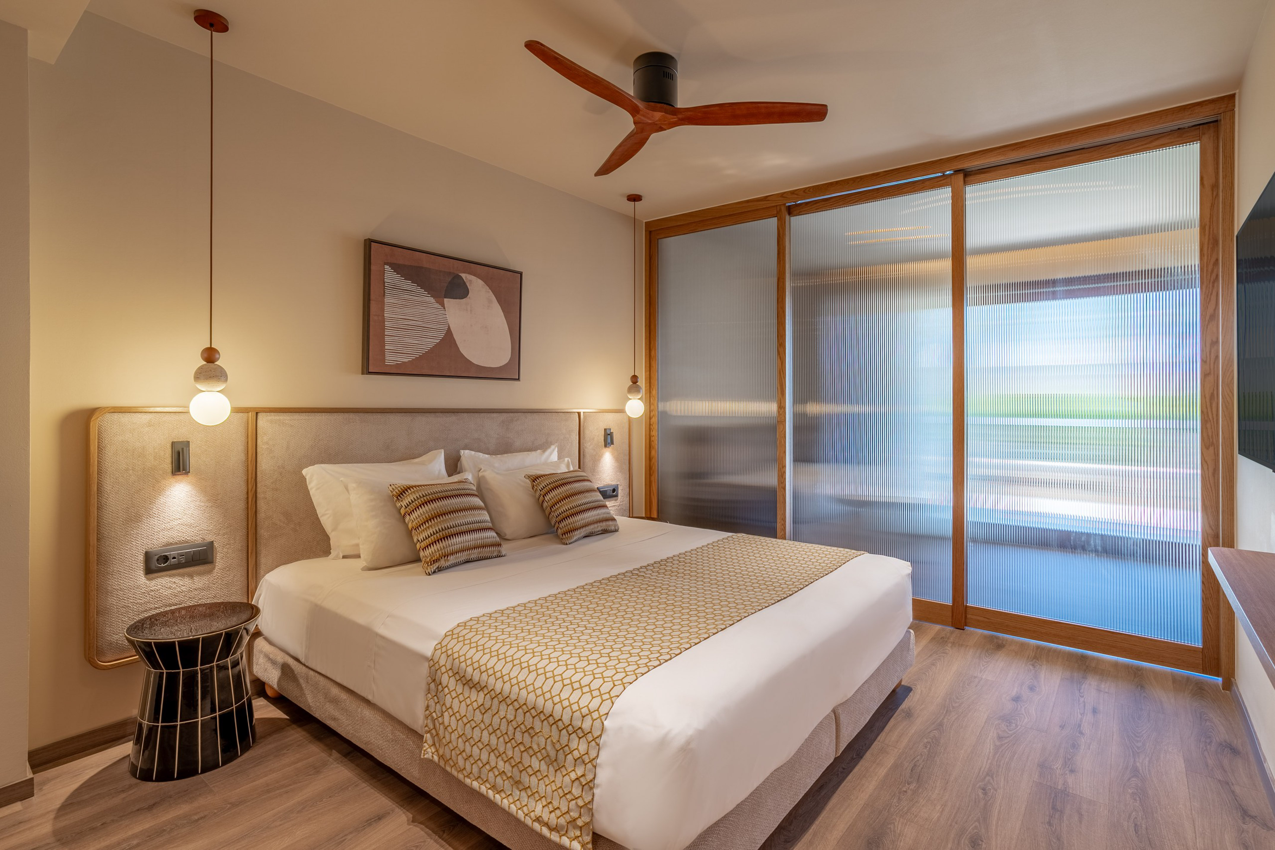 Modern hotel room with king bed, wooden ceiling fan, and fluted glass doors.