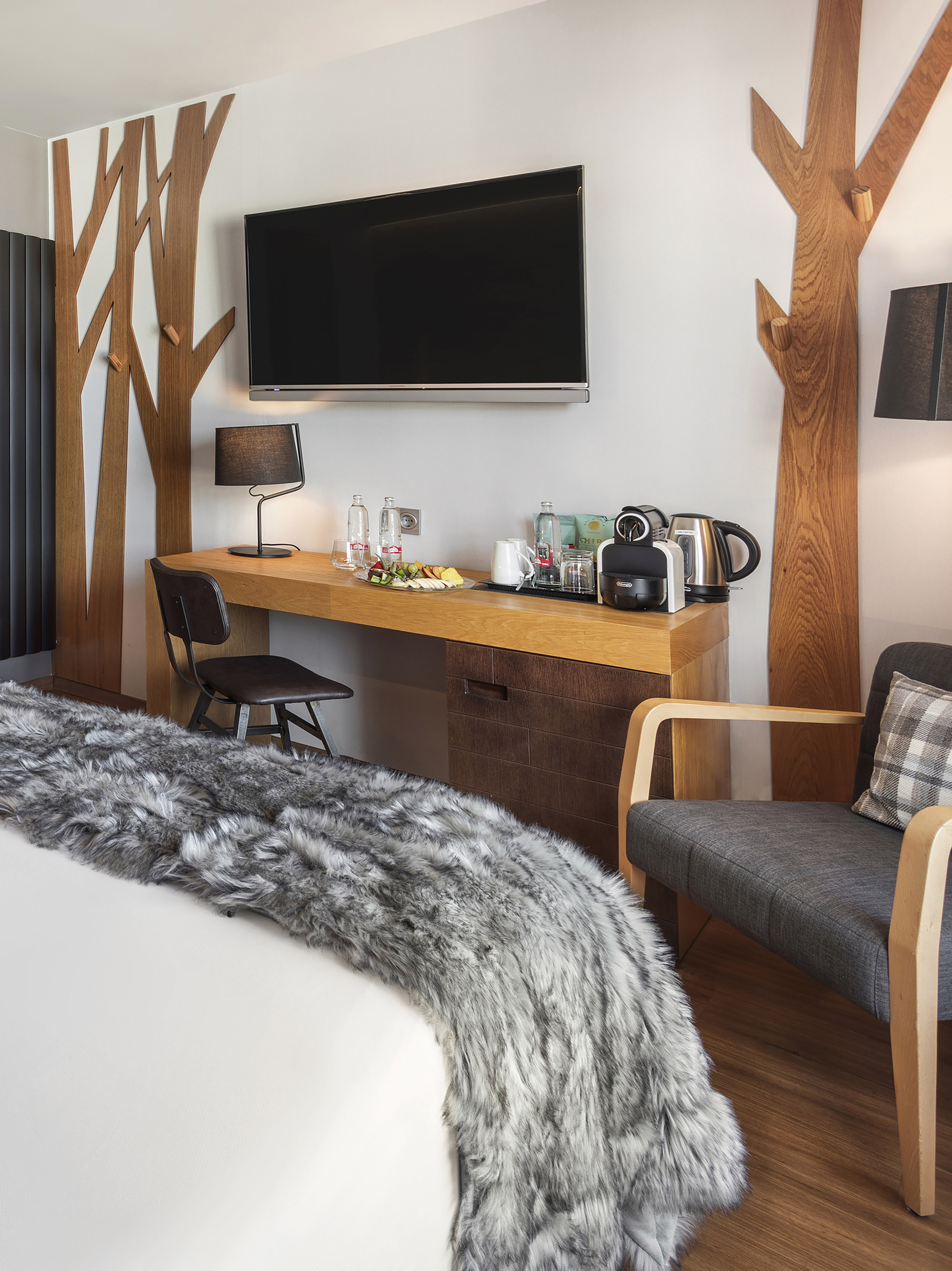 Modern hotel room with wood tree decor, TV, desk, coffee station, and gray faux fur throw.
