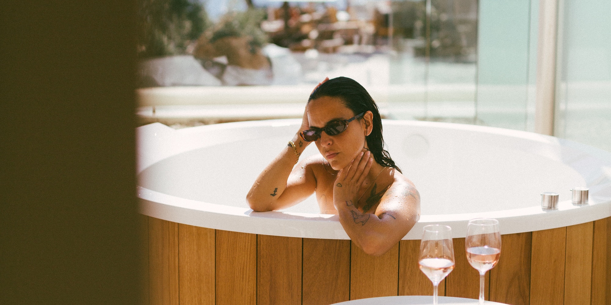 Woman in wooden hot tub with sunglasses and rosé wine outdoors.