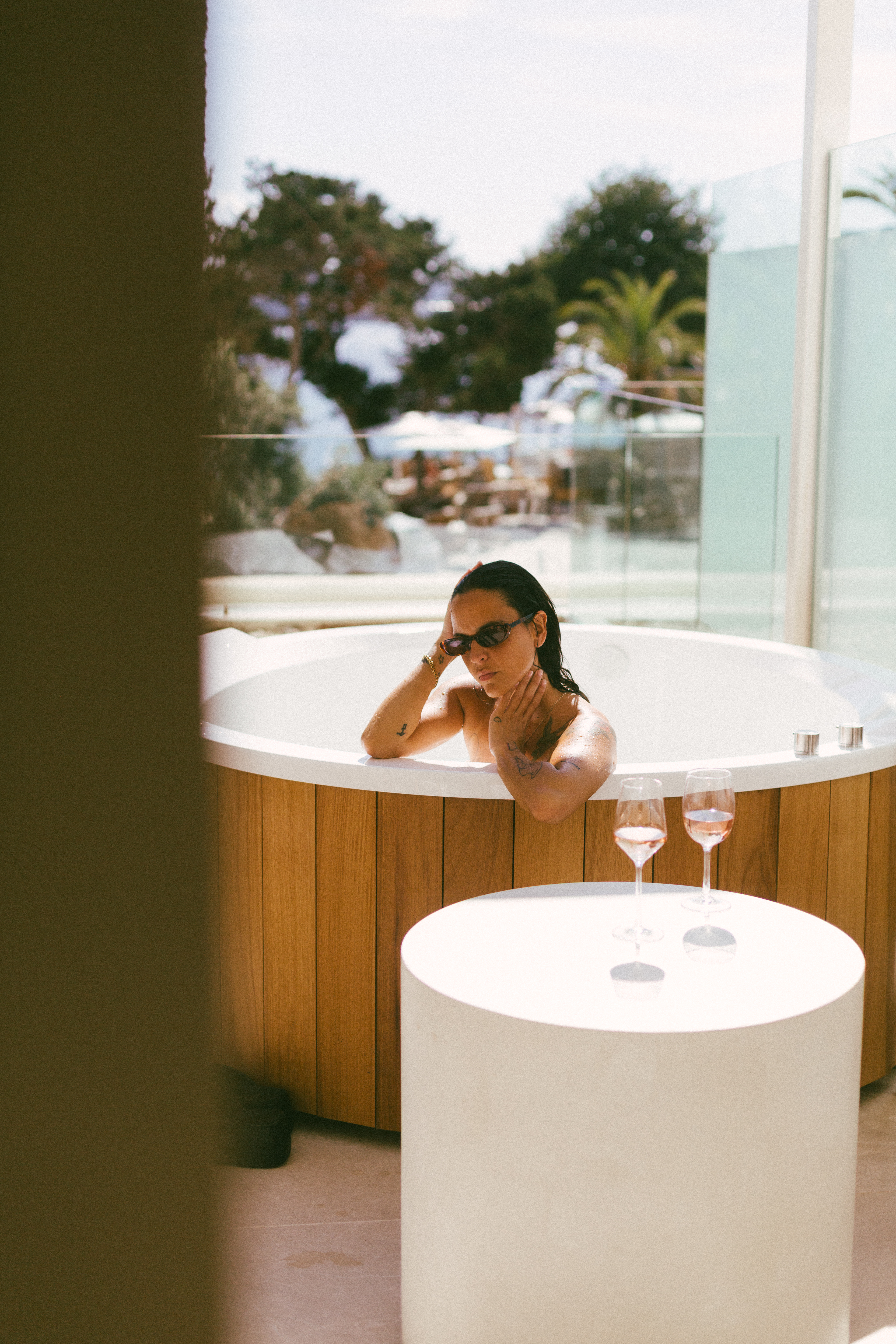 Woman in wooden hot tub with sunglasses and rosé wine outdoors.