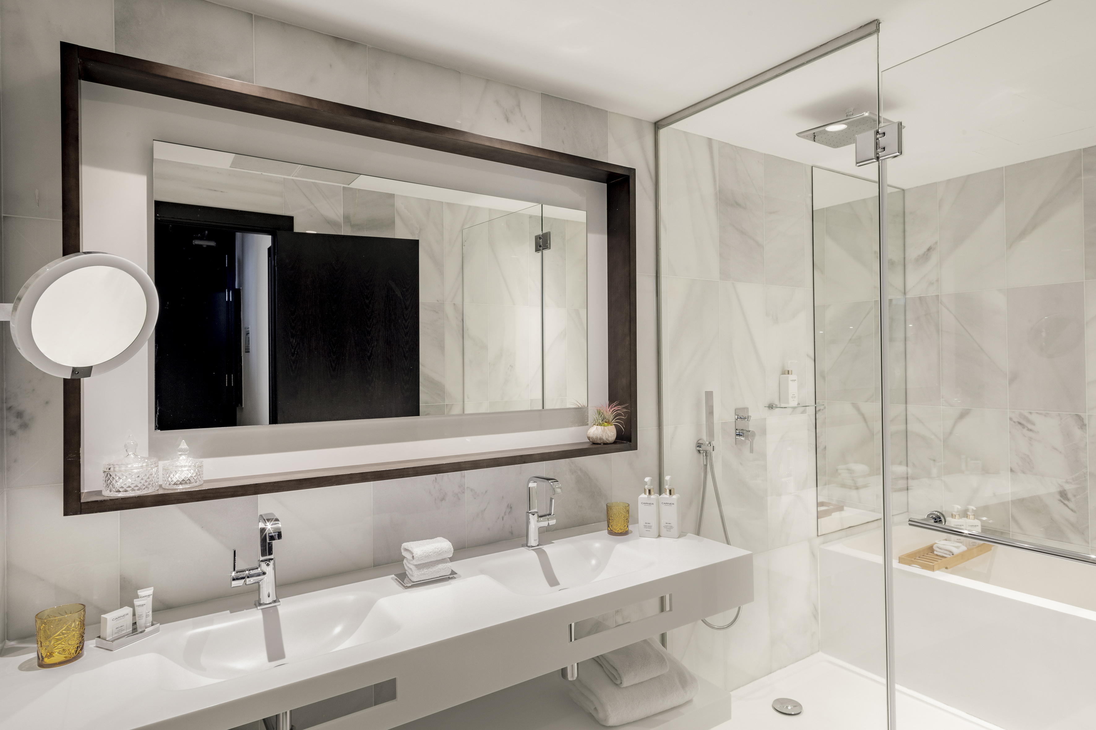 Modern marble hotel bathroom with double vanity, large mirror, glass shower.