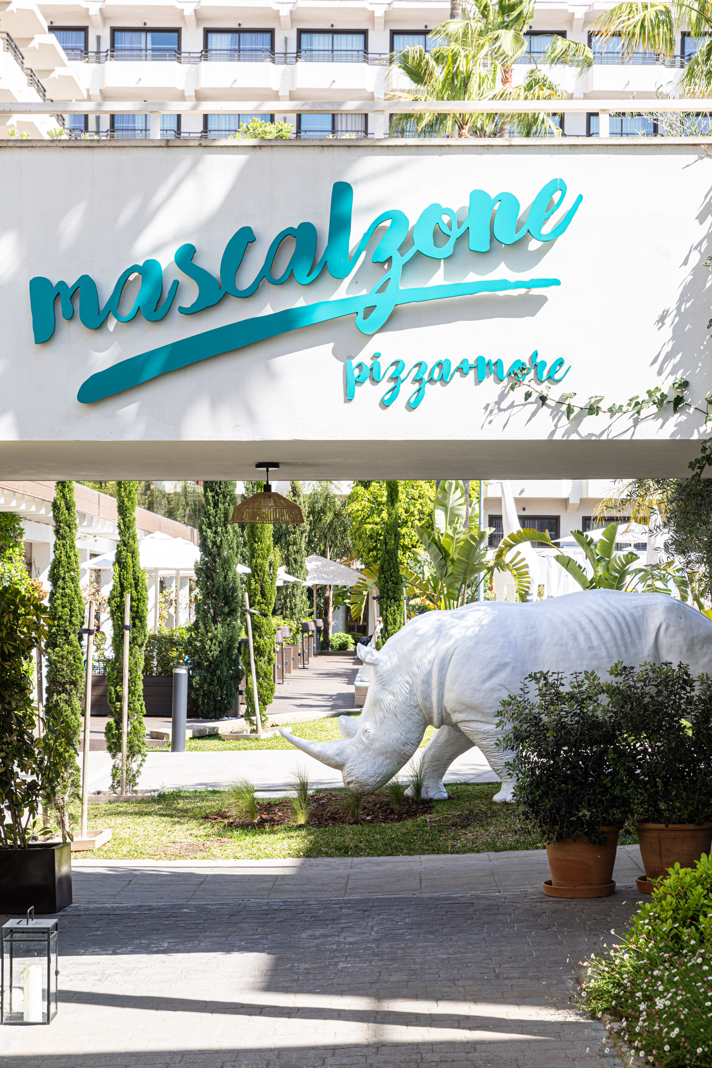 Mascalzone pizza+more sign, white rhino statue in a lush resort garden.