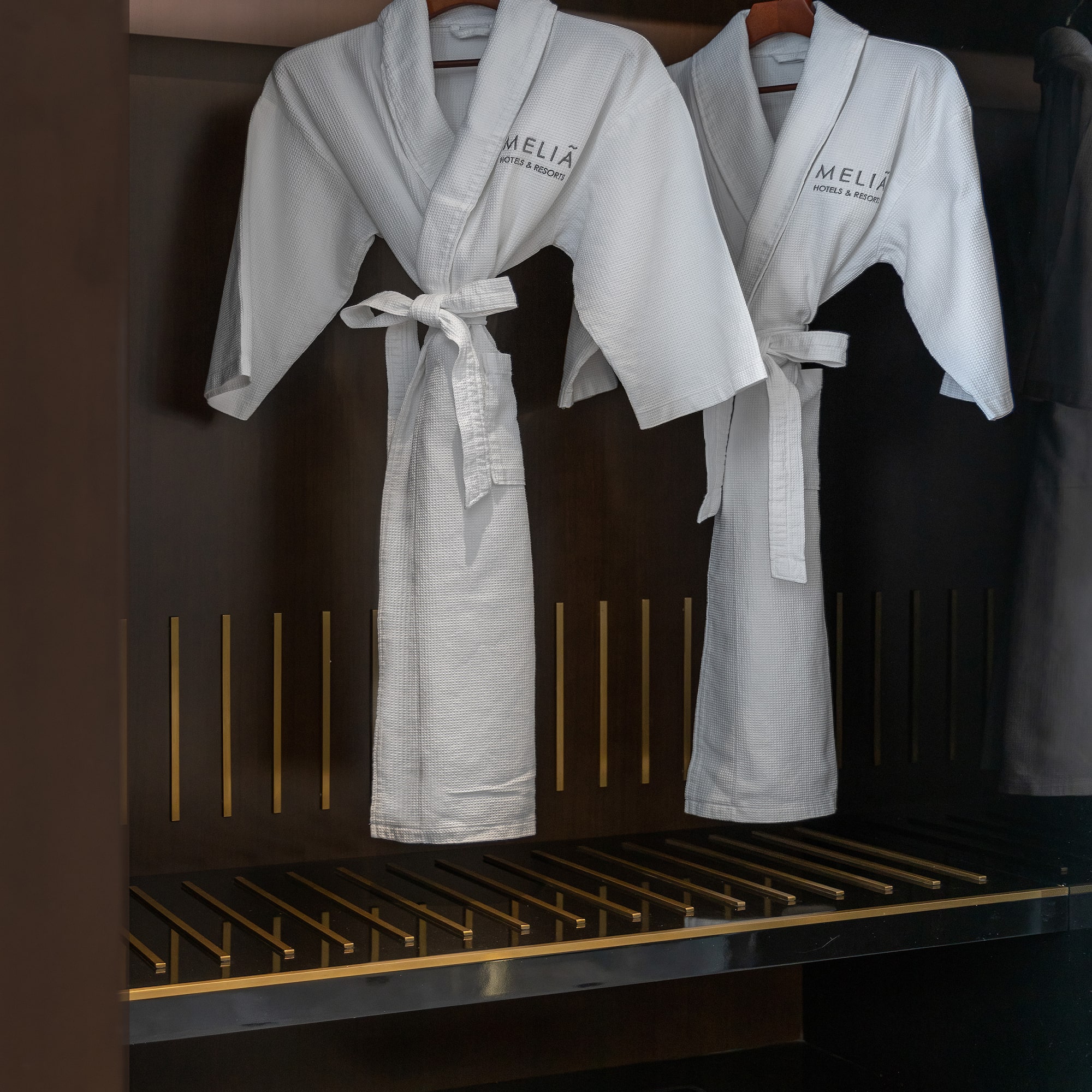 Two white Meliá hotel bathrobes and slippers in a dark wardrobe.