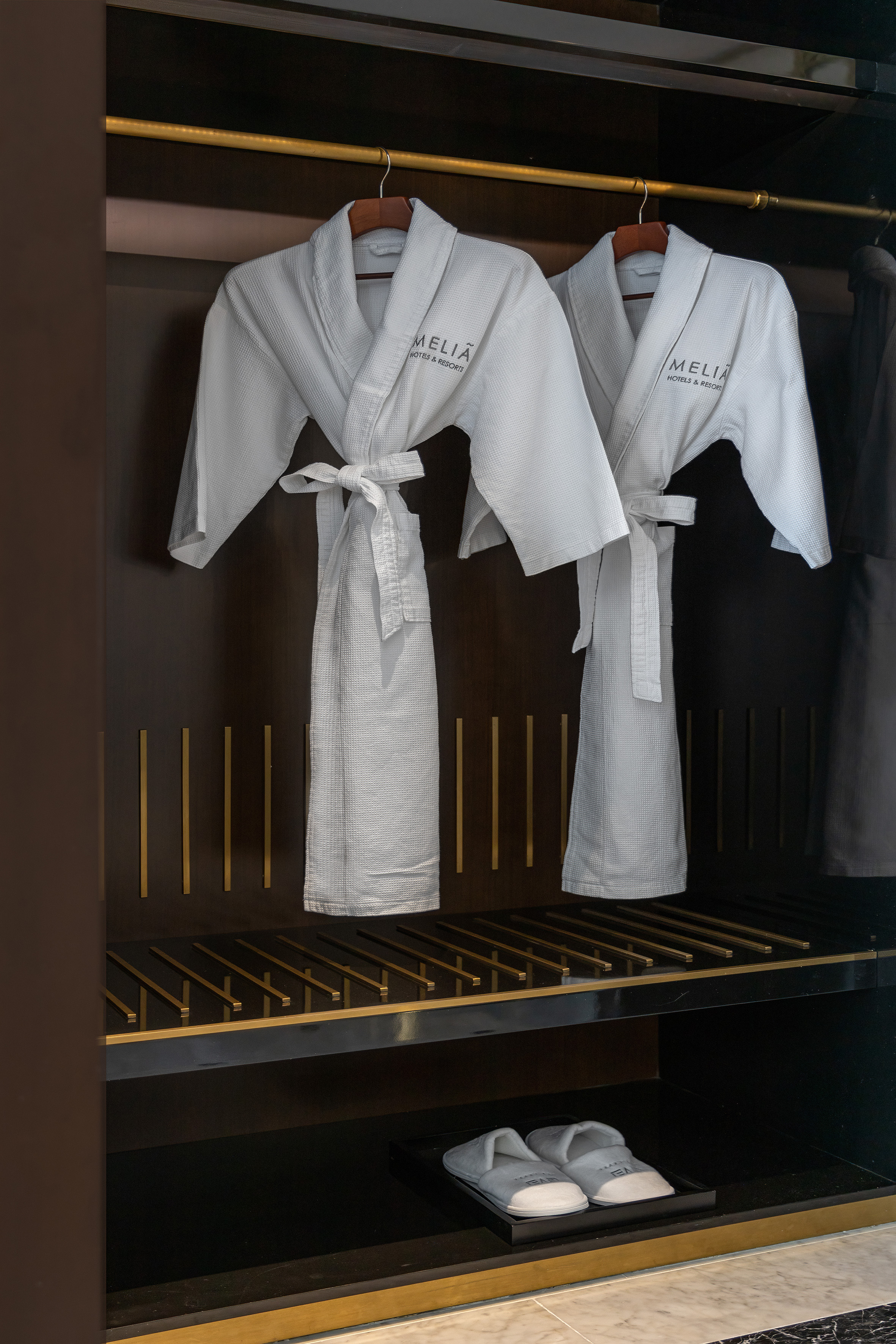 Two white Meliá hotel bathrobes and slippers in a dark wardrobe.