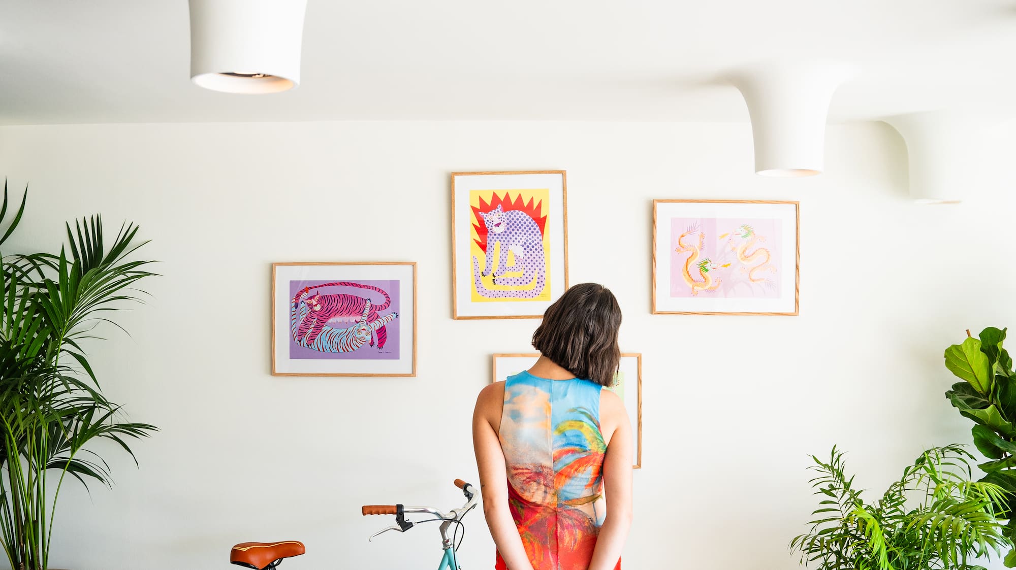 Woman in colorful dress by teal bike, colorful art prints, plants, black panther statue.