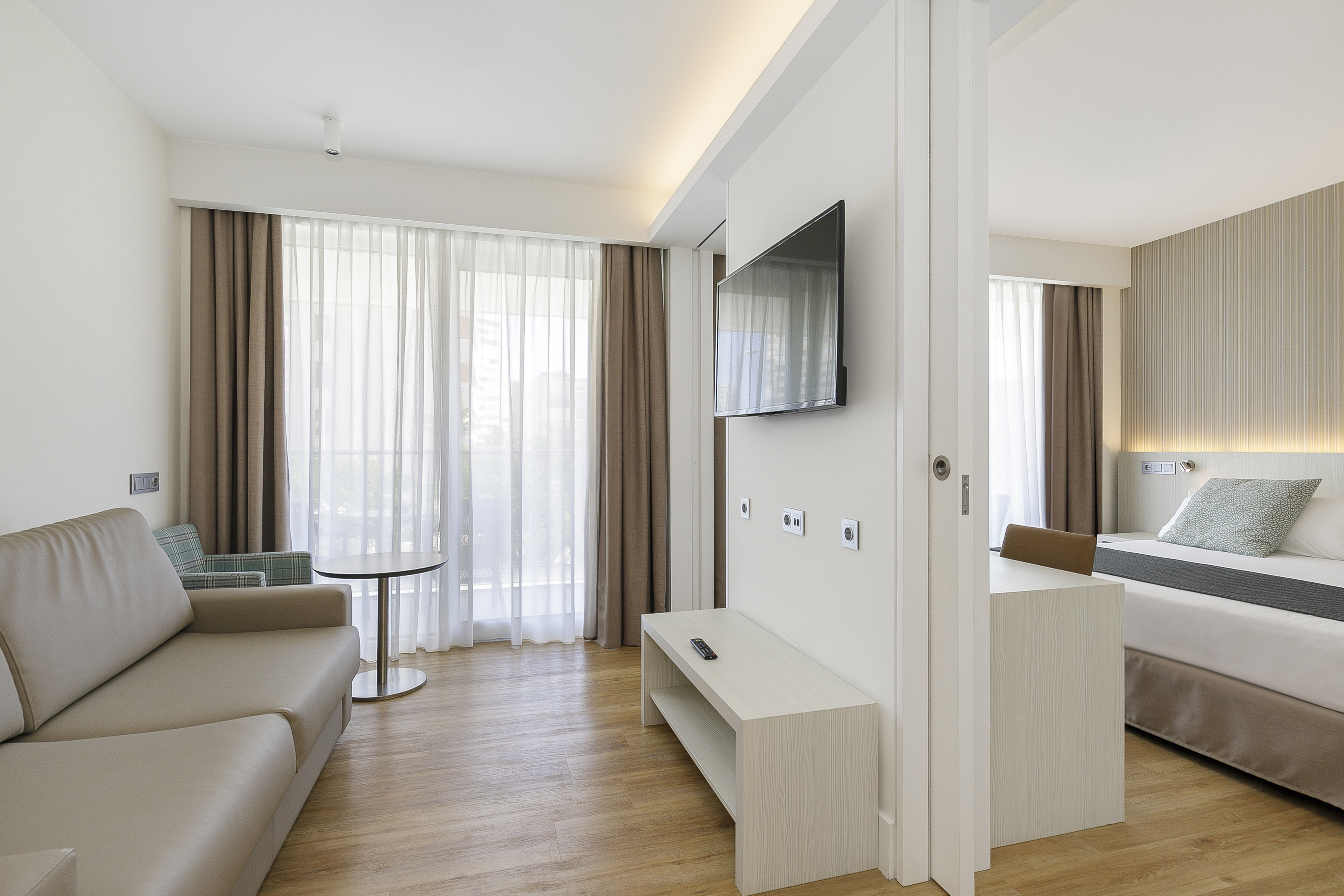 Bright, modern hotel suite with living area, sofa, TV, and contemporary bedroom.