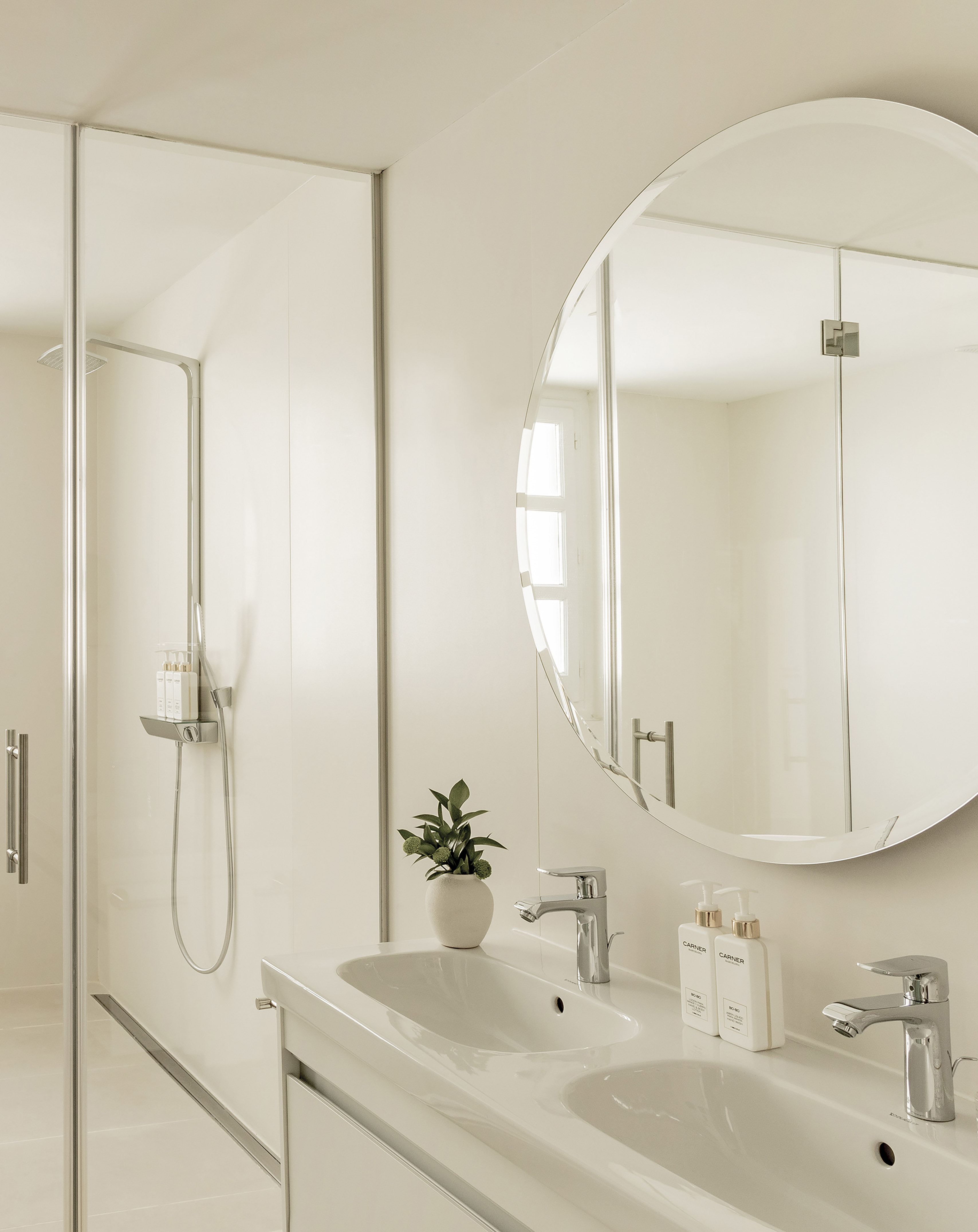 Minimalist white bathroom with double sinks, round mirror, and glass shower.