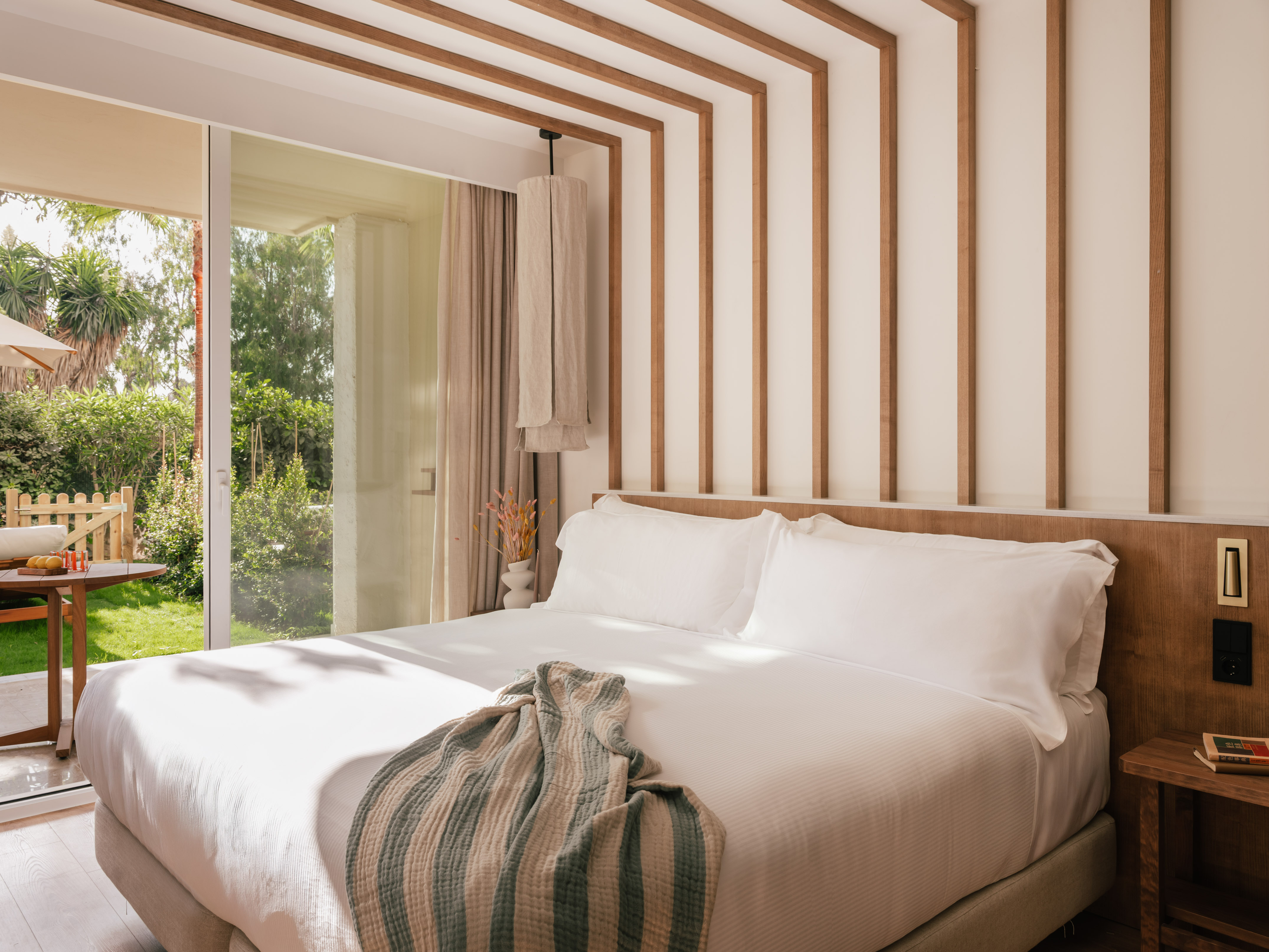 Modern hotel bedroom with natural wood accents, white bed, and garden view patio.