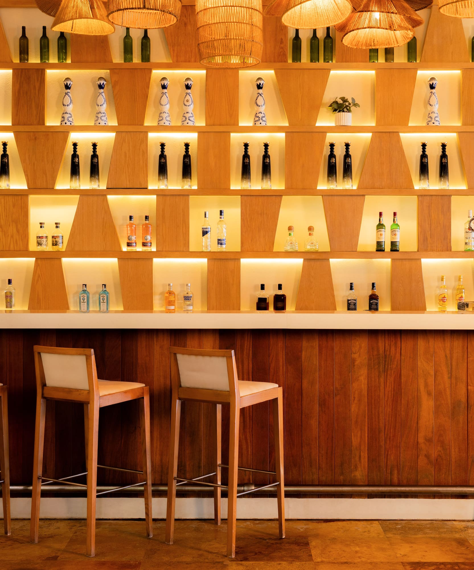 Illuminated wooden bar with liquor bottles on shelves, three stools, and rattan lights.