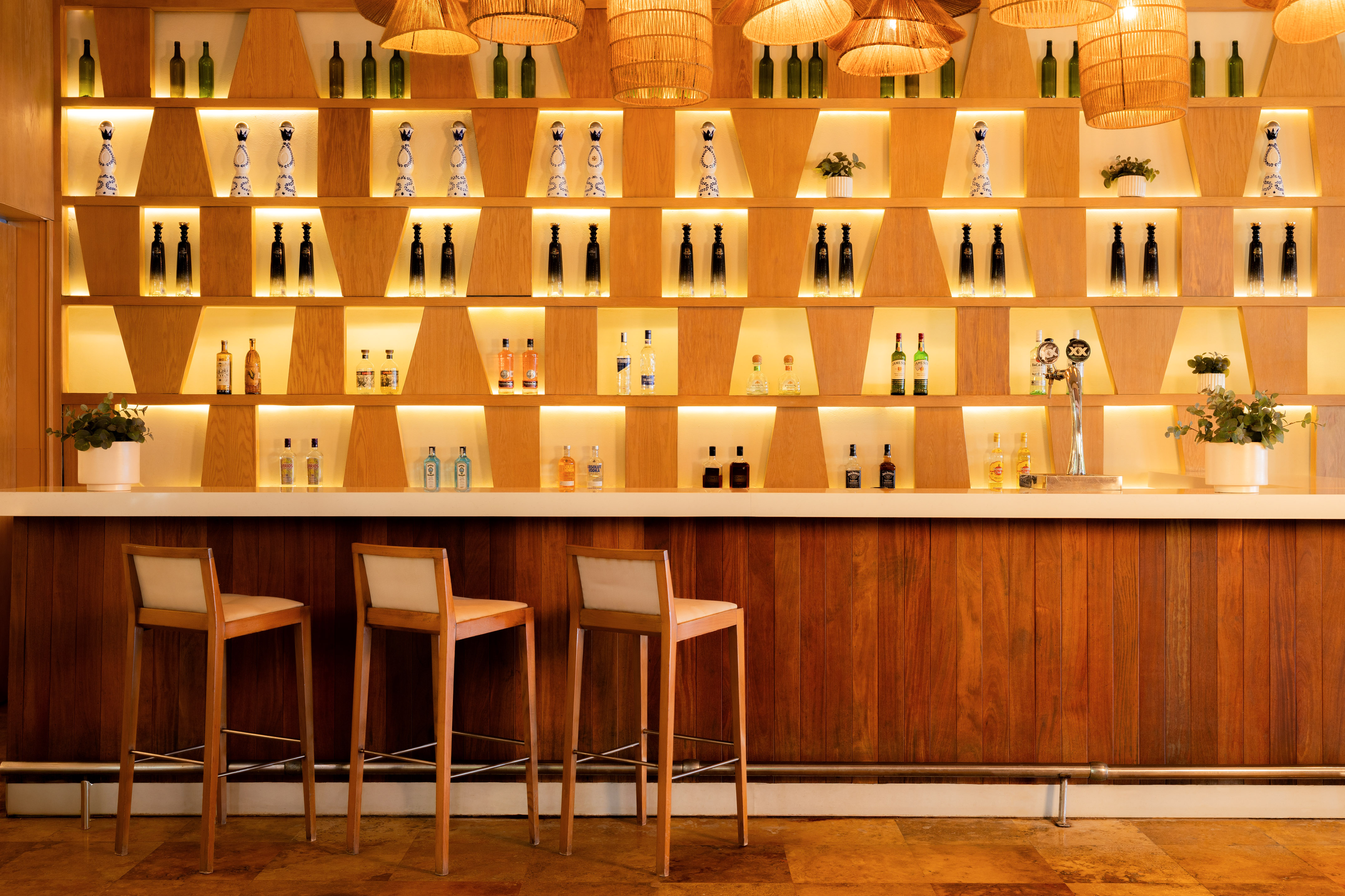 Illuminated wooden bar with liquor bottles on shelves, three stools, and rattan lights.
