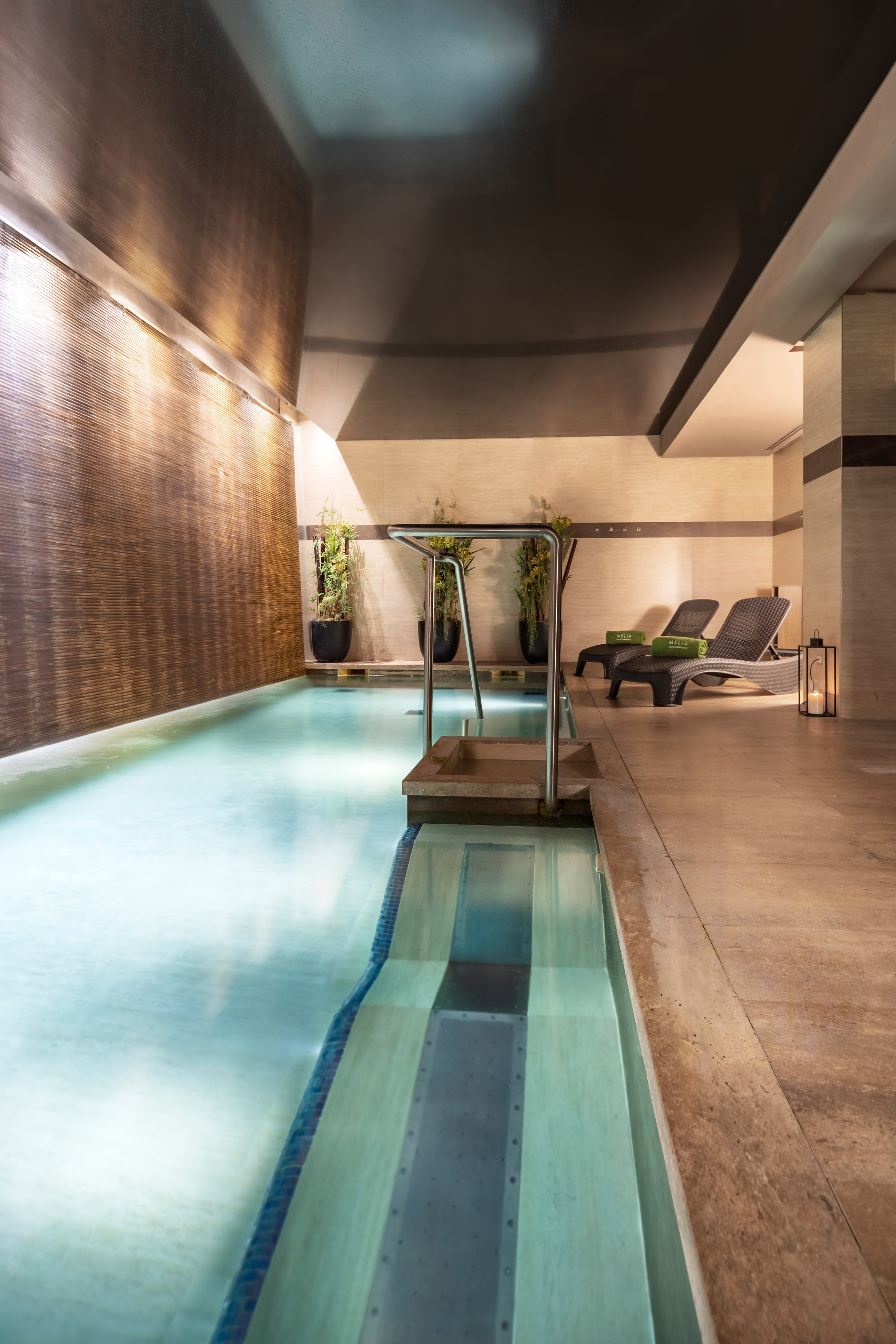 Modern indoor spa pool with textured water wall, lounge chairs, and plants. - YHI Wellness - Meliá Genova, Genua