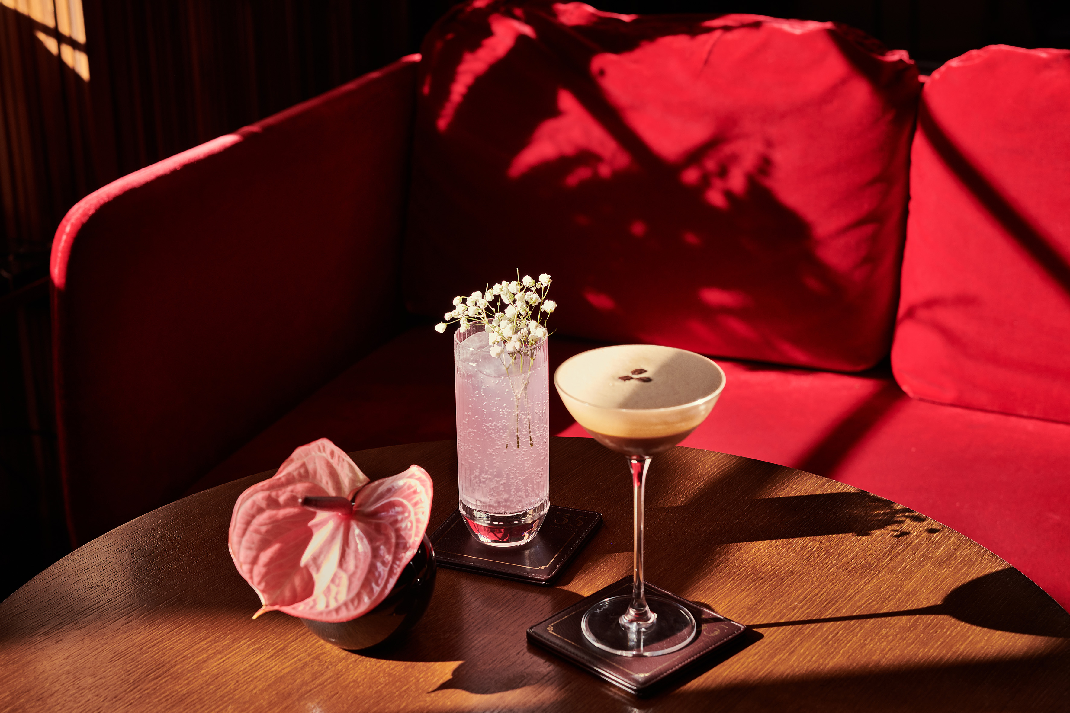 Espresso martini, pink cocktail, anthurium on wooden table, red lounge sofa.