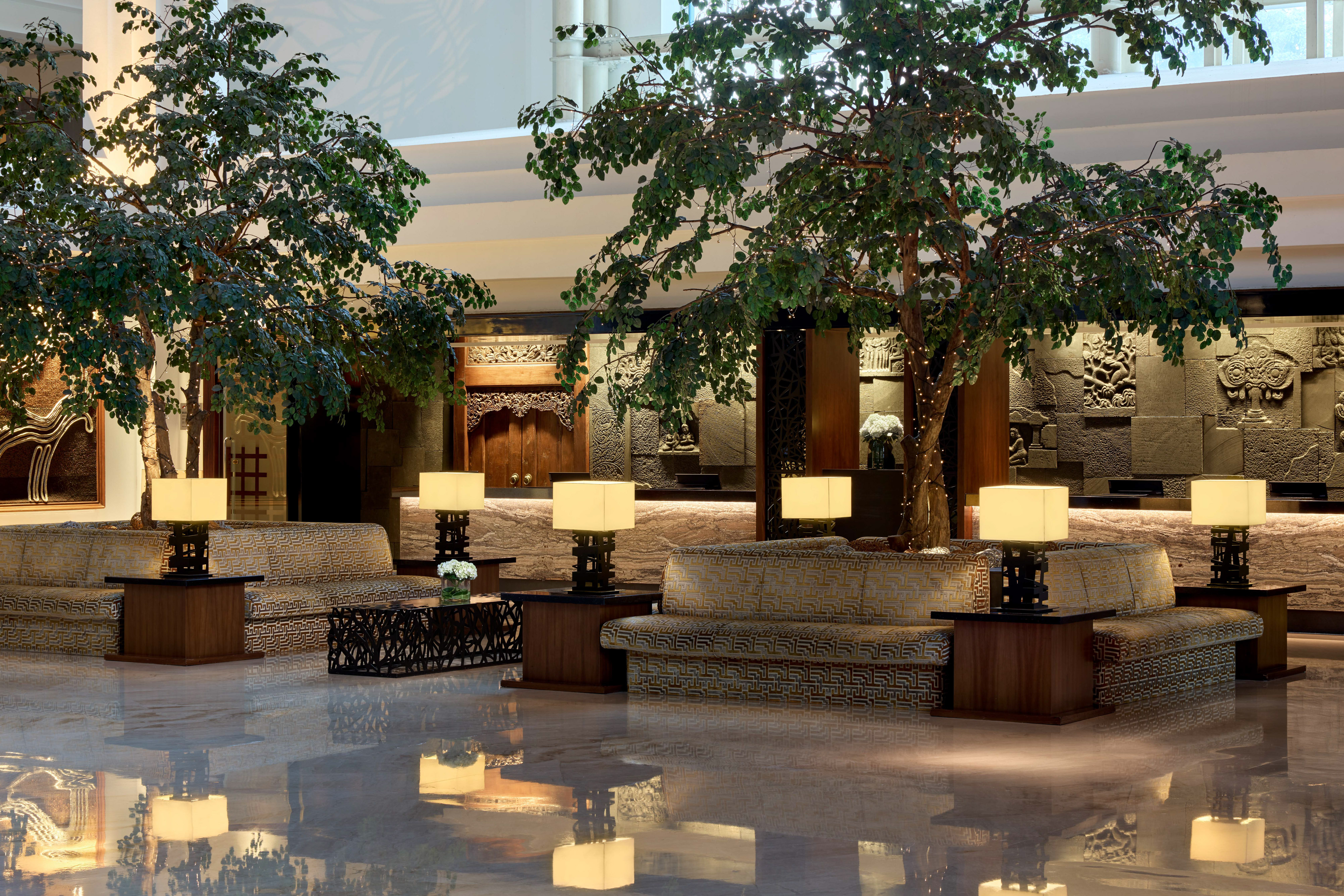 Elegant hotel lobby with large tropical trees, patterned sofas, and Asian-inspired decor.