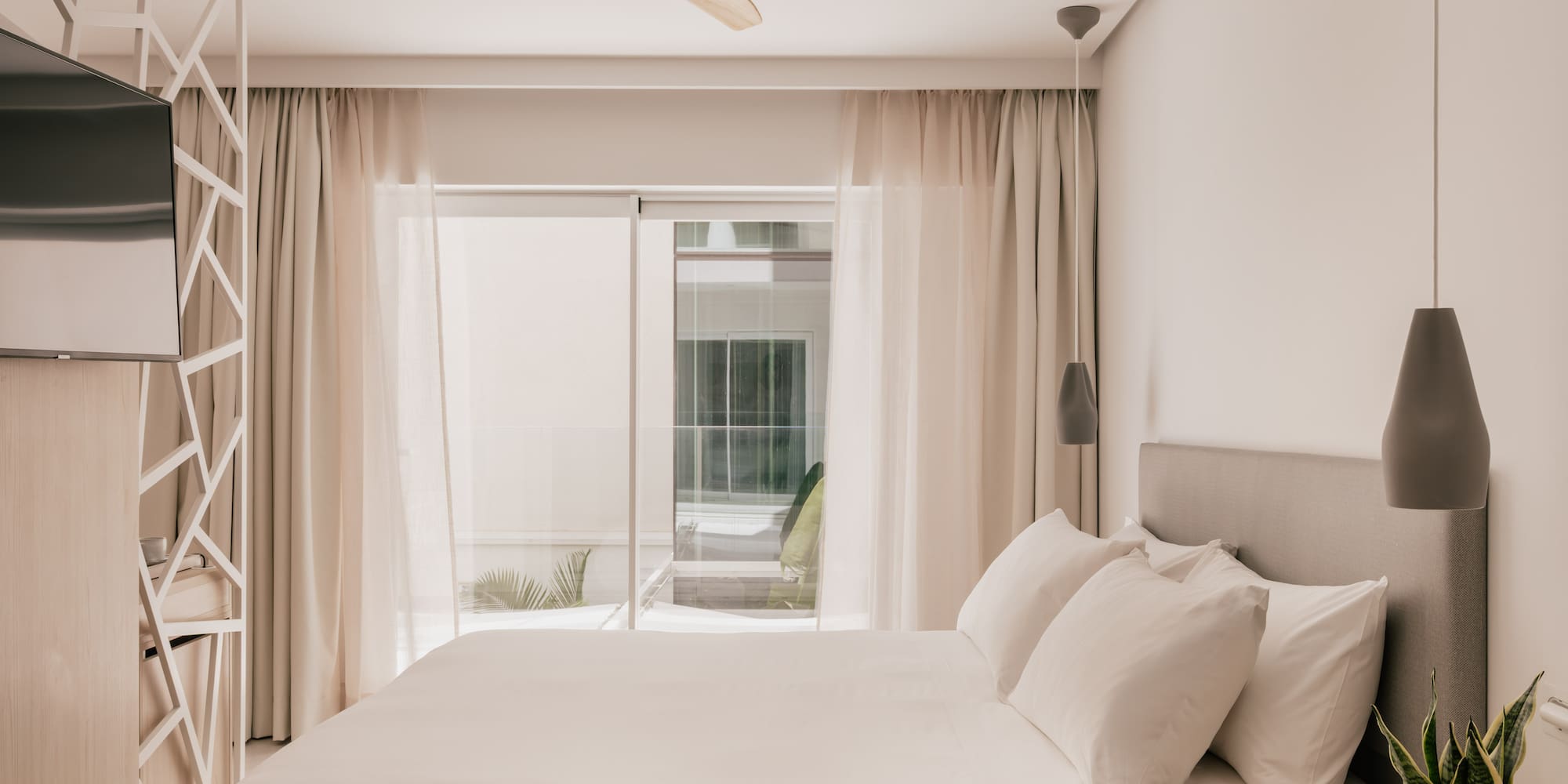 Modern hotel bedroom with white bed, wooden ceiling fan, and bright window.