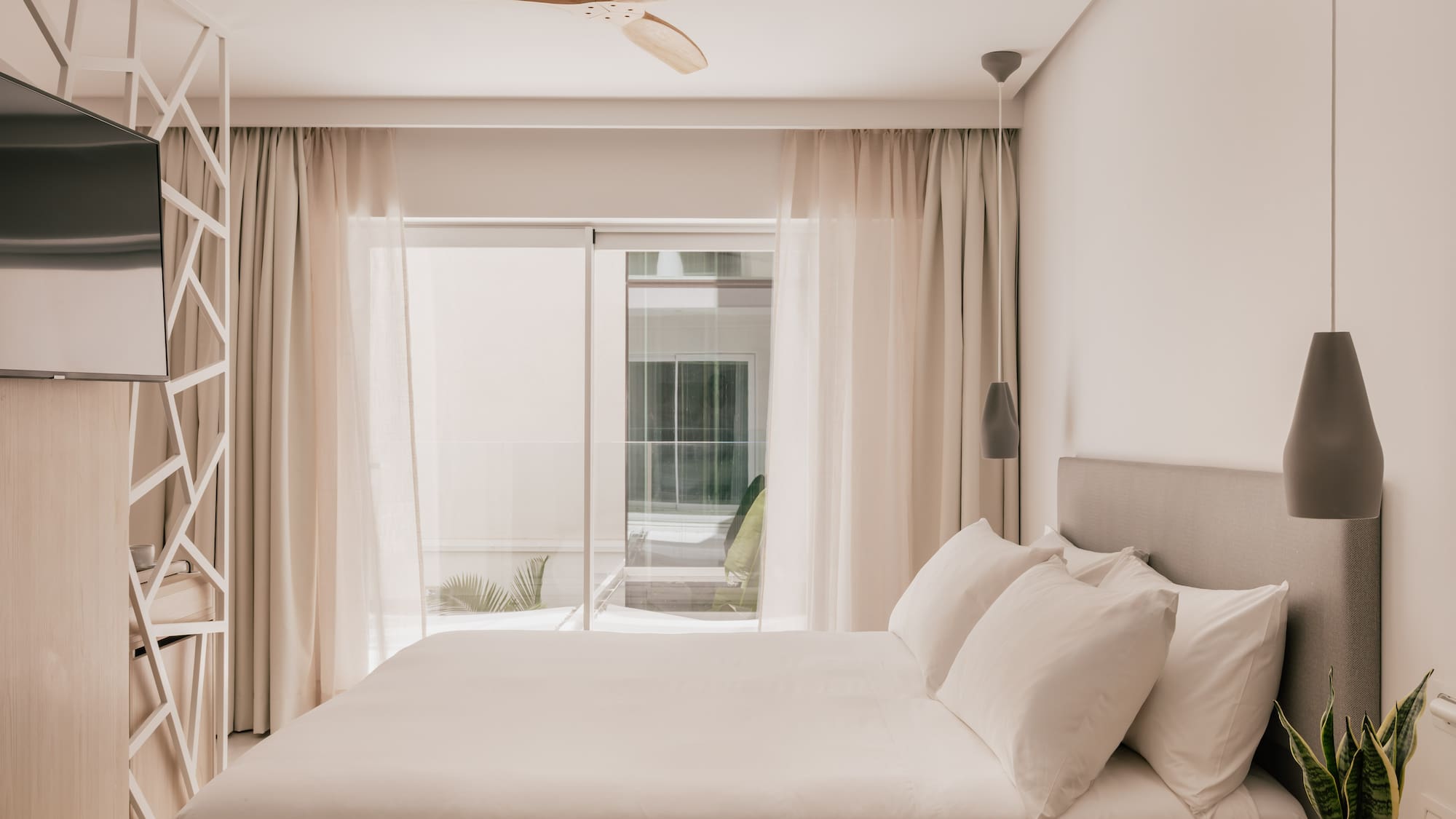 Modern hotel bedroom with white bed, wooden ceiling fan, and bright window.
