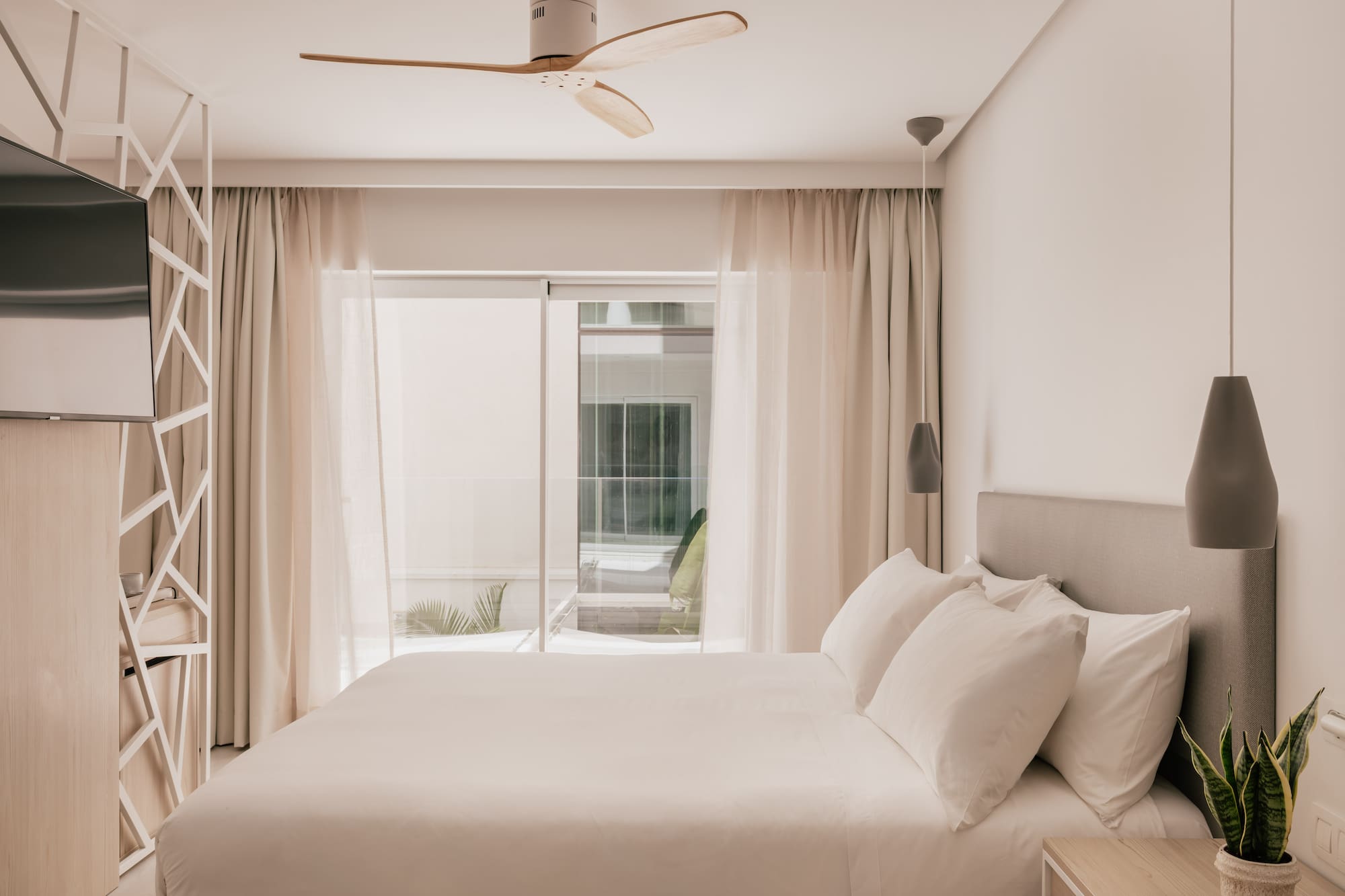 Modern hotel bedroom with white bed, wooden ceiling fan, and bright window.