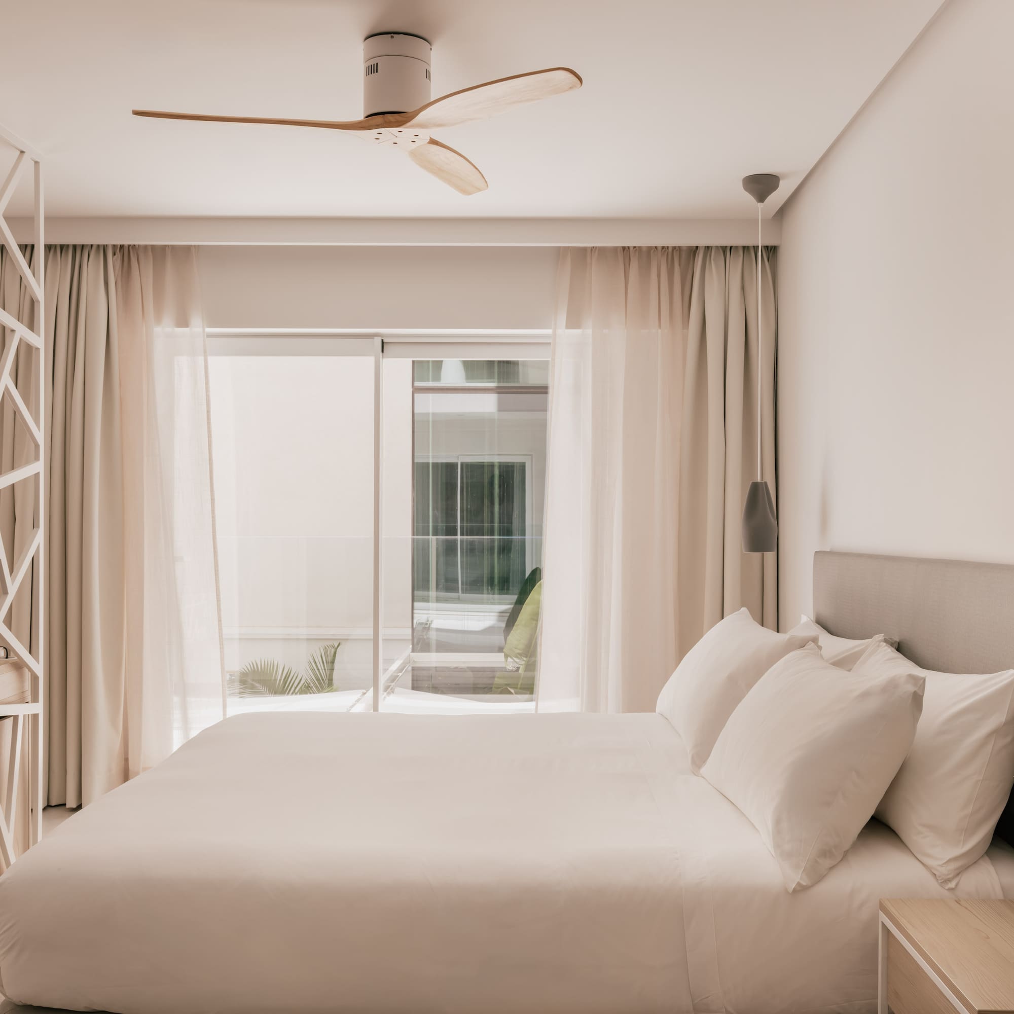 Modern hotel bedroom with white bed, wooden ceiling fan, and bright window.