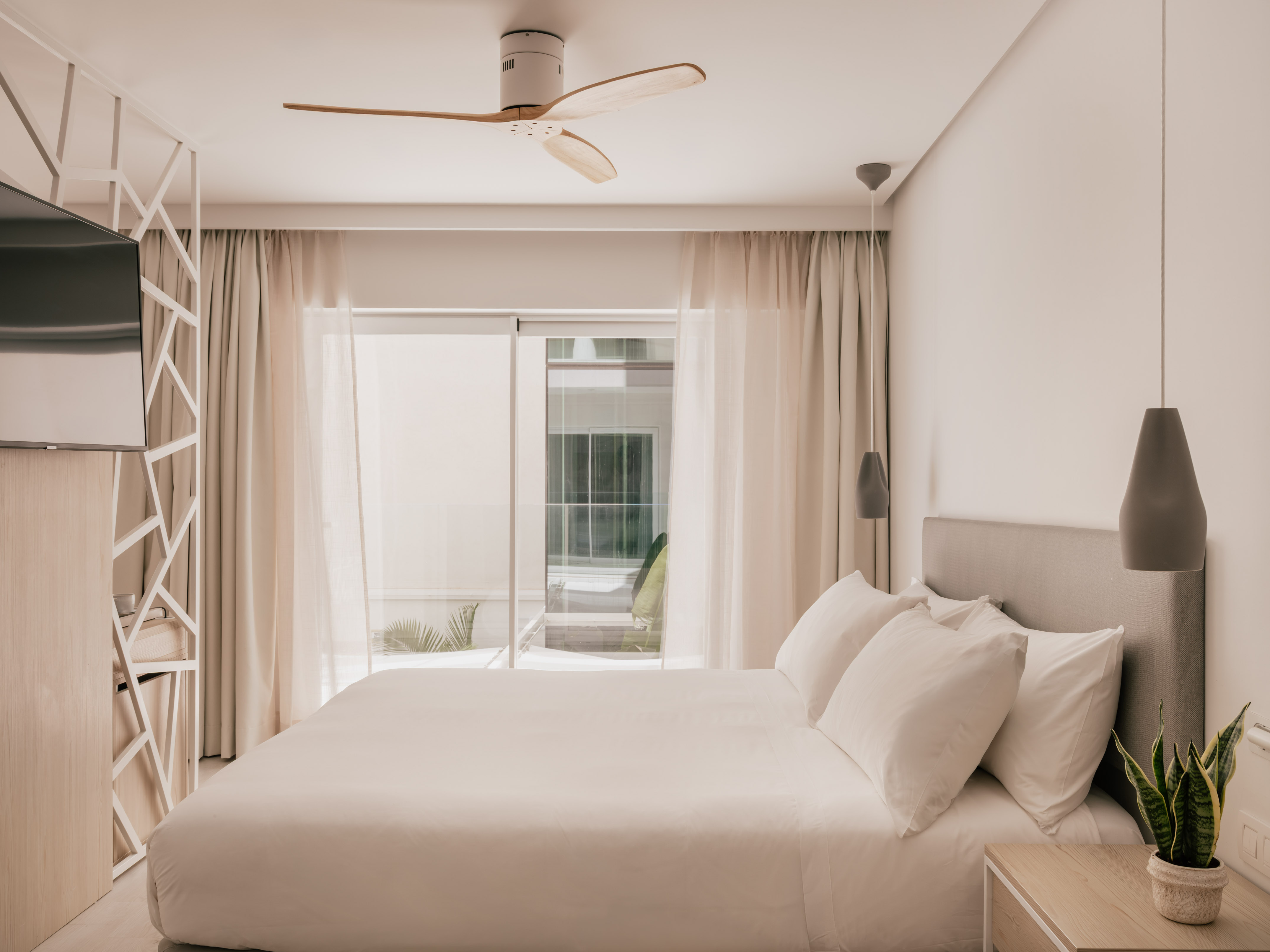 Modern hotel bedroom with white bed, wooden ceiling fan, and bright window.