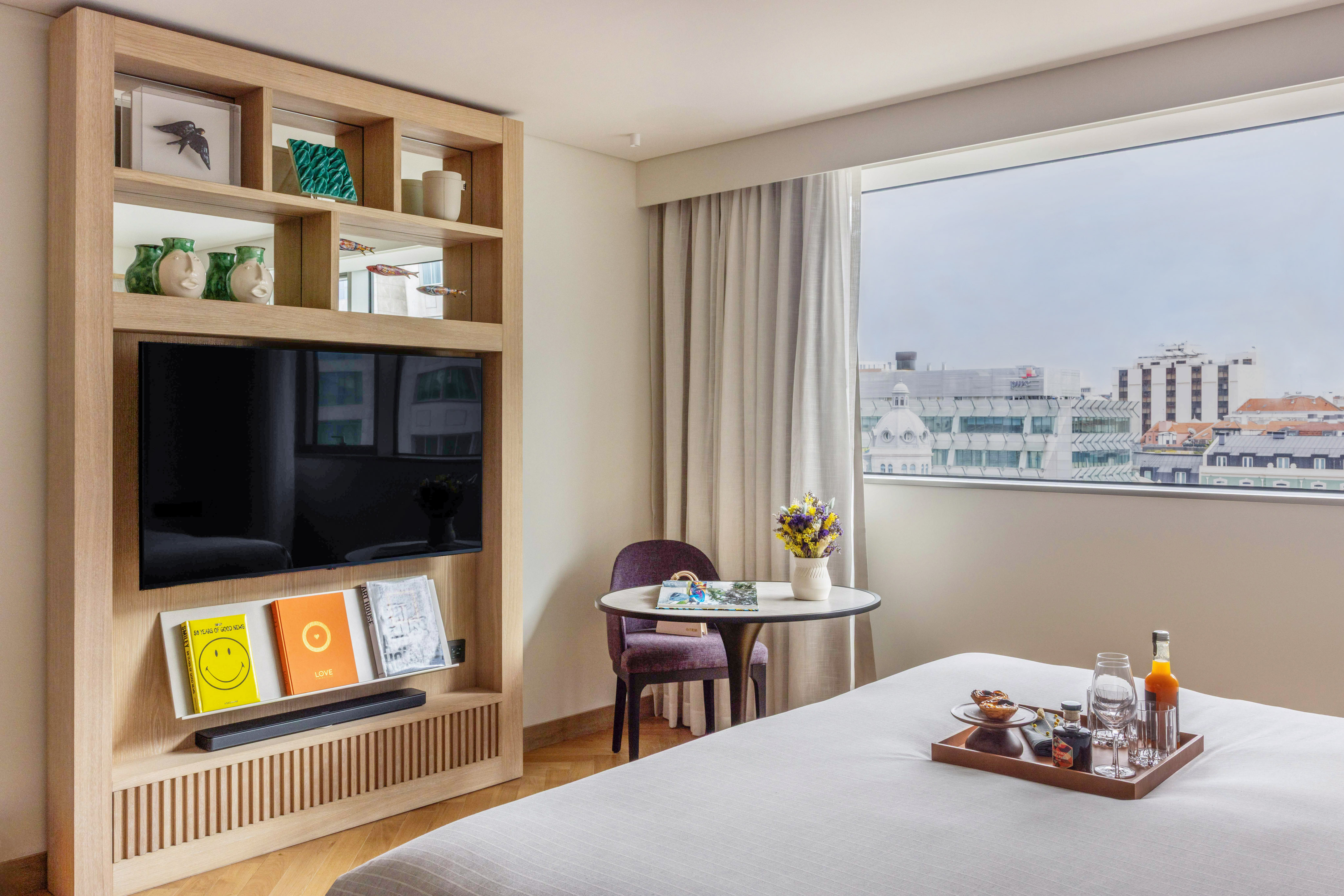 Modern hotel room with city view, breakfast on bed, and integrated TV unit.